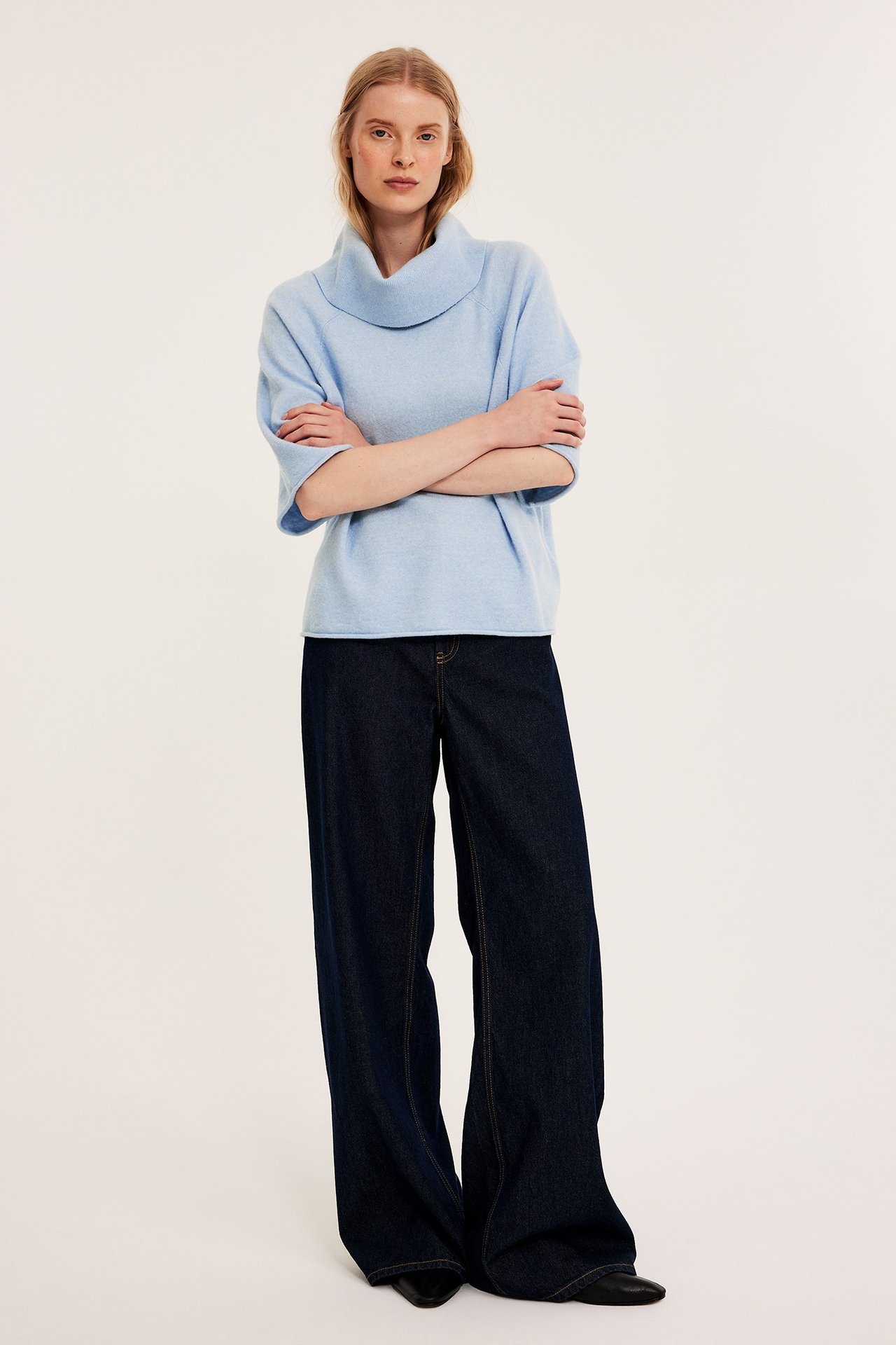 Widok z przodu: kobieta w jasnoniebieskim melanżowym swetrze z golfem, ciemnych szerokich dżinsach i czarnych butach.