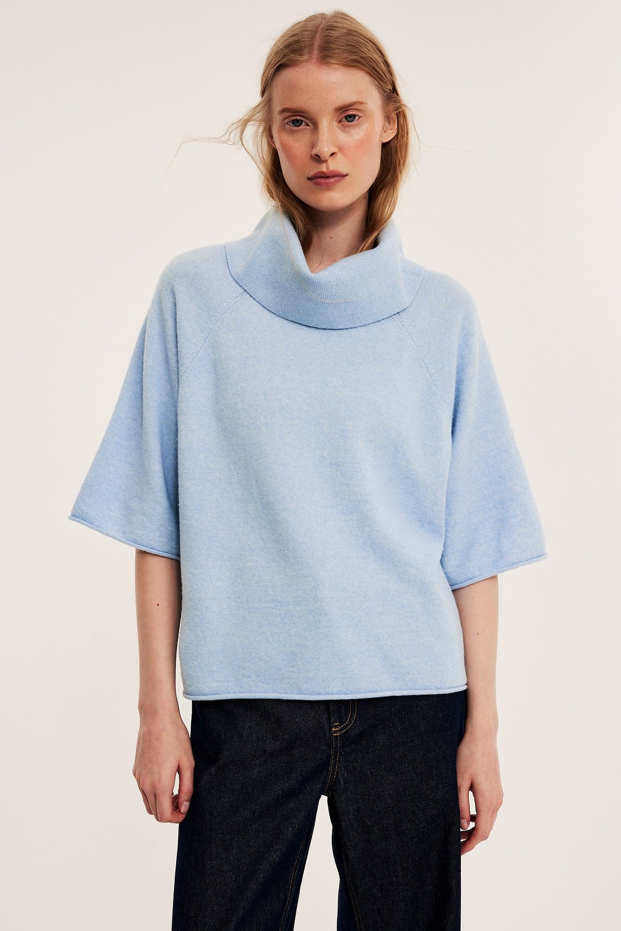 Widok z przodu: kobieta w jasnoniebieskim melanżowym swetrze z golfem, krótkie rękawy, ciemne dżinsy.