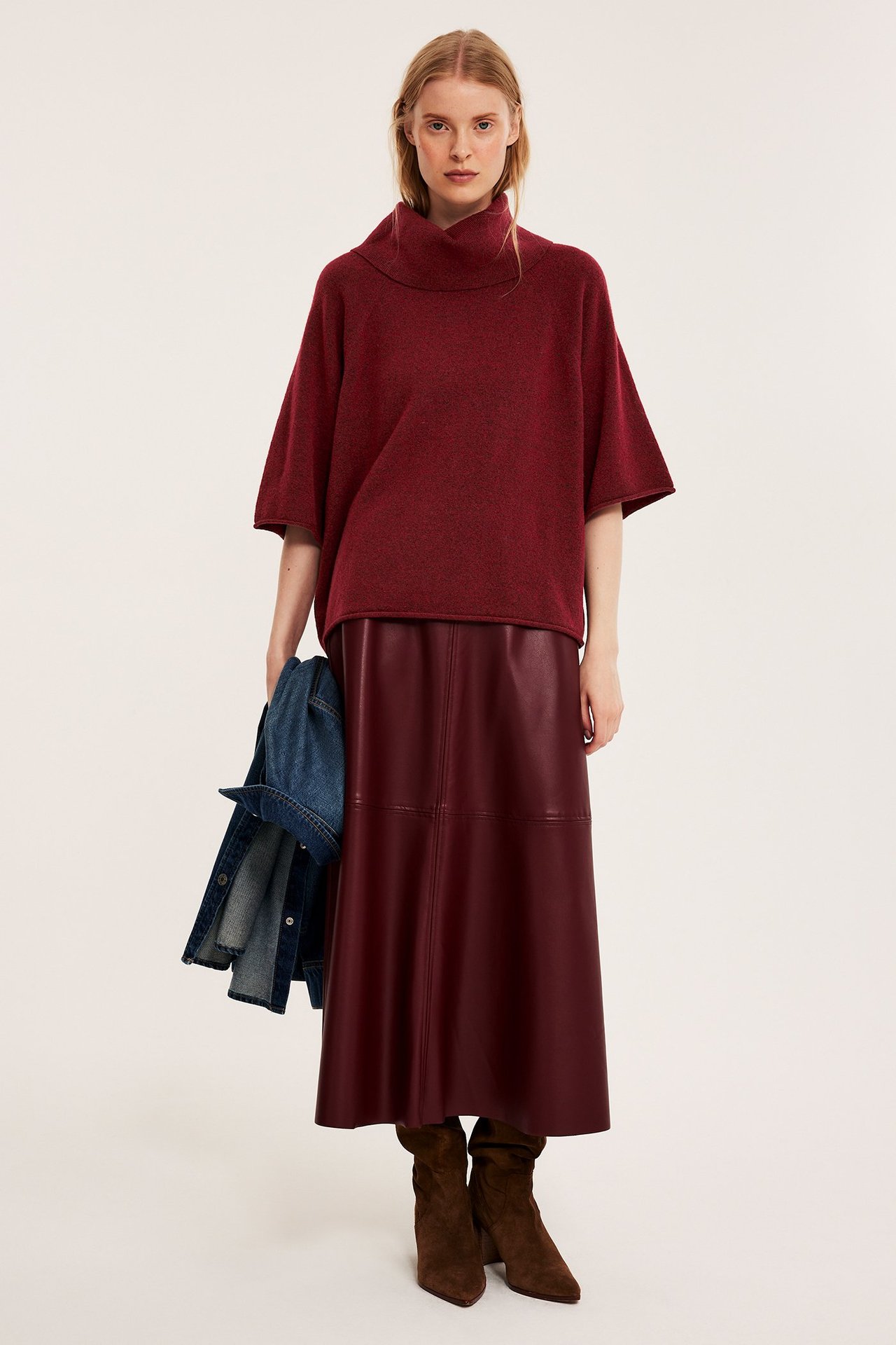 Front view: Kvinna i rödmelerad stickad polotröja, mörkröd kjol, bruna stövlar, håller blå jeansjacka.