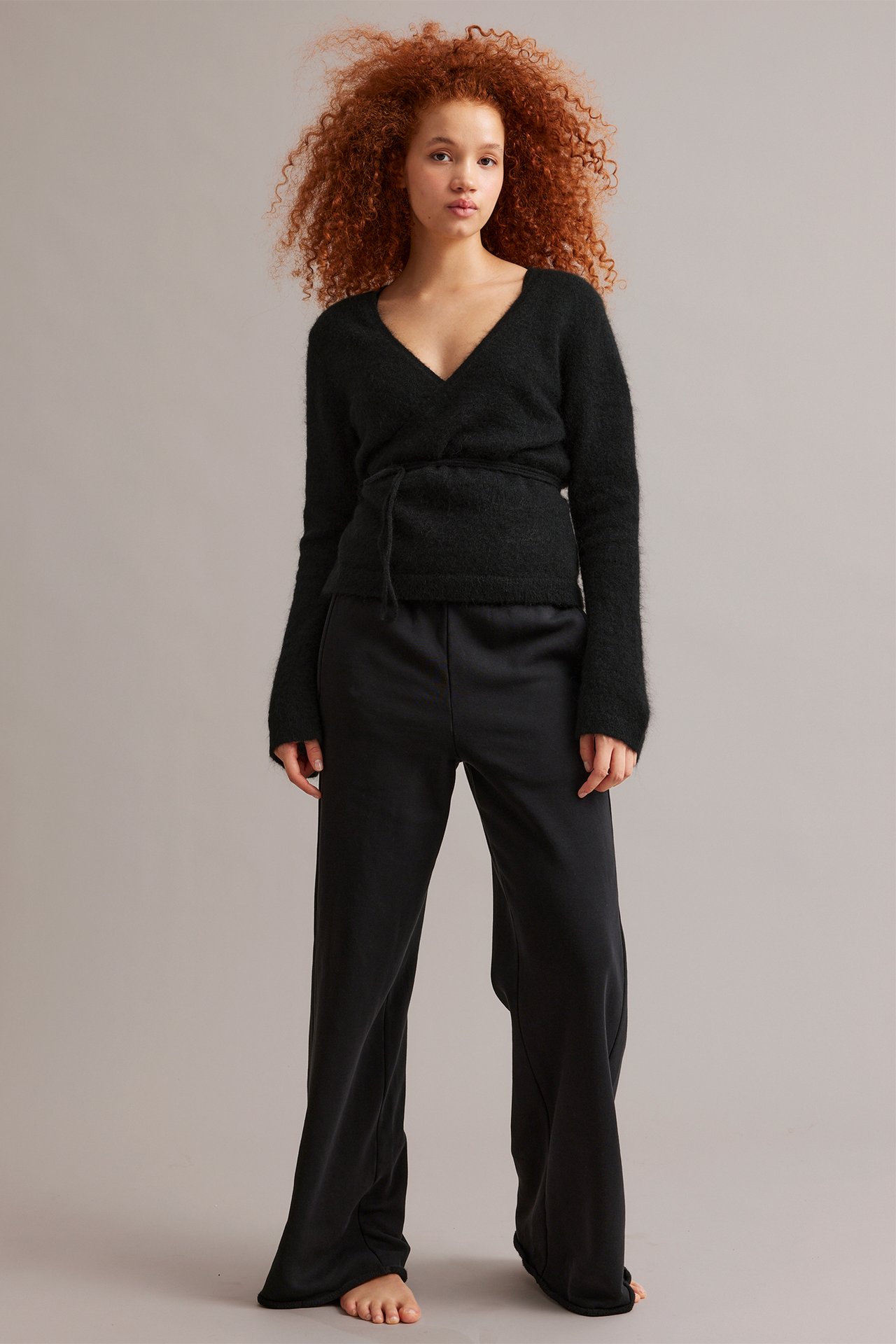 Edestäpäin, nainen yllään musta villasekoite kietaisuneuletakki leveillä hihoilla ja mustat leveälahkeiset housut.