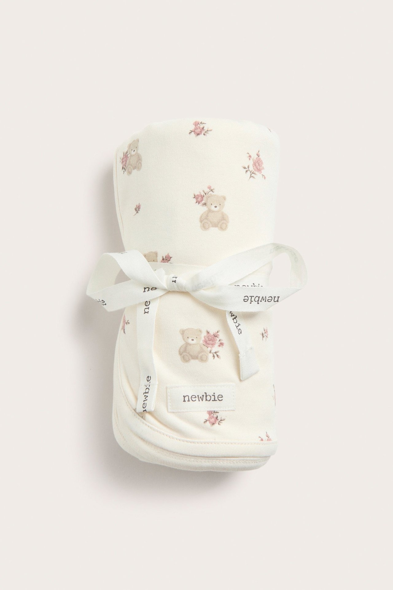 Edestä: Taitettu luonnonvalkoinen puuvillavauvanpeitto, jossa karhuja ja vaaleanpunaisia ruusuja, sidottu valkoisella nauhalla.