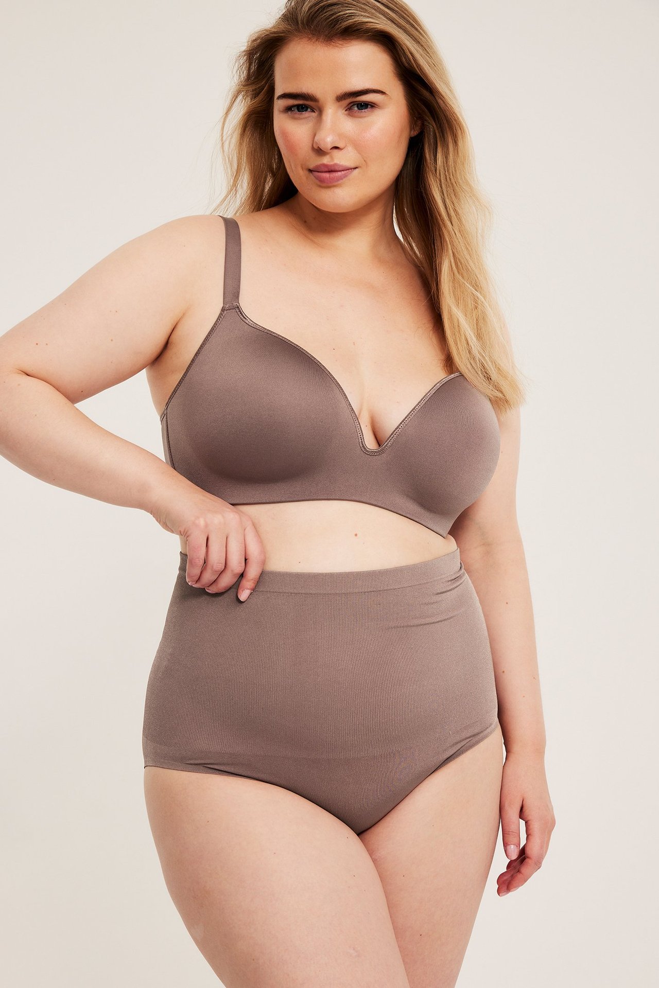 Naisen etukuva yllään korkeavyötäröiset, taupe saumattomat alushousut ja yhteensopivat rintaliivit.