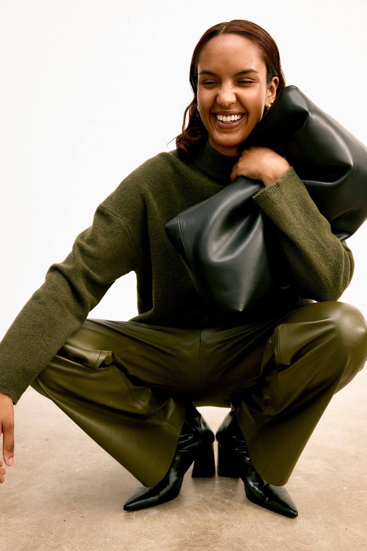 Edestä: Nainen vihreissä tekonahkahousuissa, vihreässä neulepuserossa, mustissa saappaissa, pitäen mustaa laukkua.