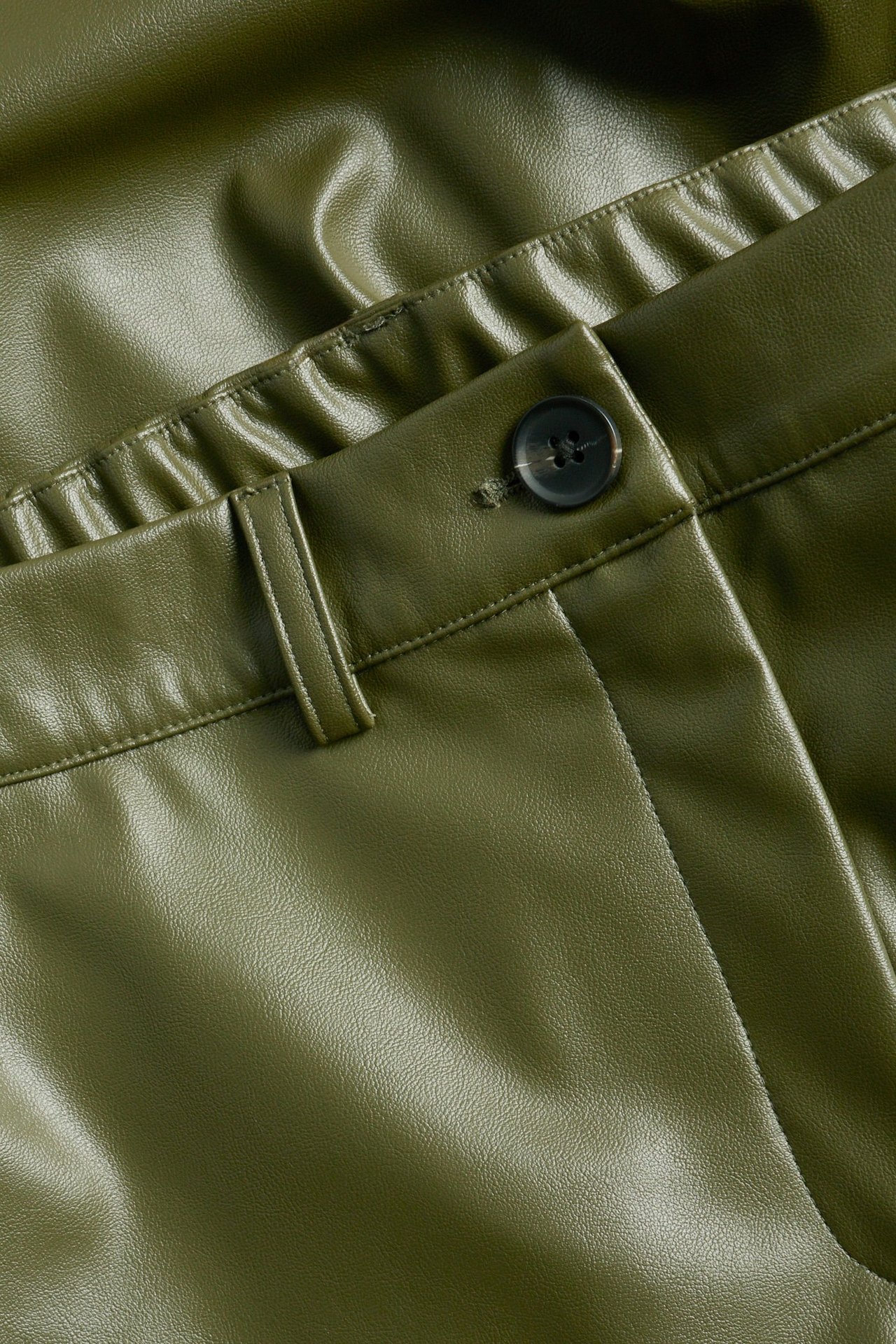 Zbliżenie zielonych spodni ze sztucznej skóry z zapięciem na guzik, szlufkami i elastycznym pasem z tyłu.