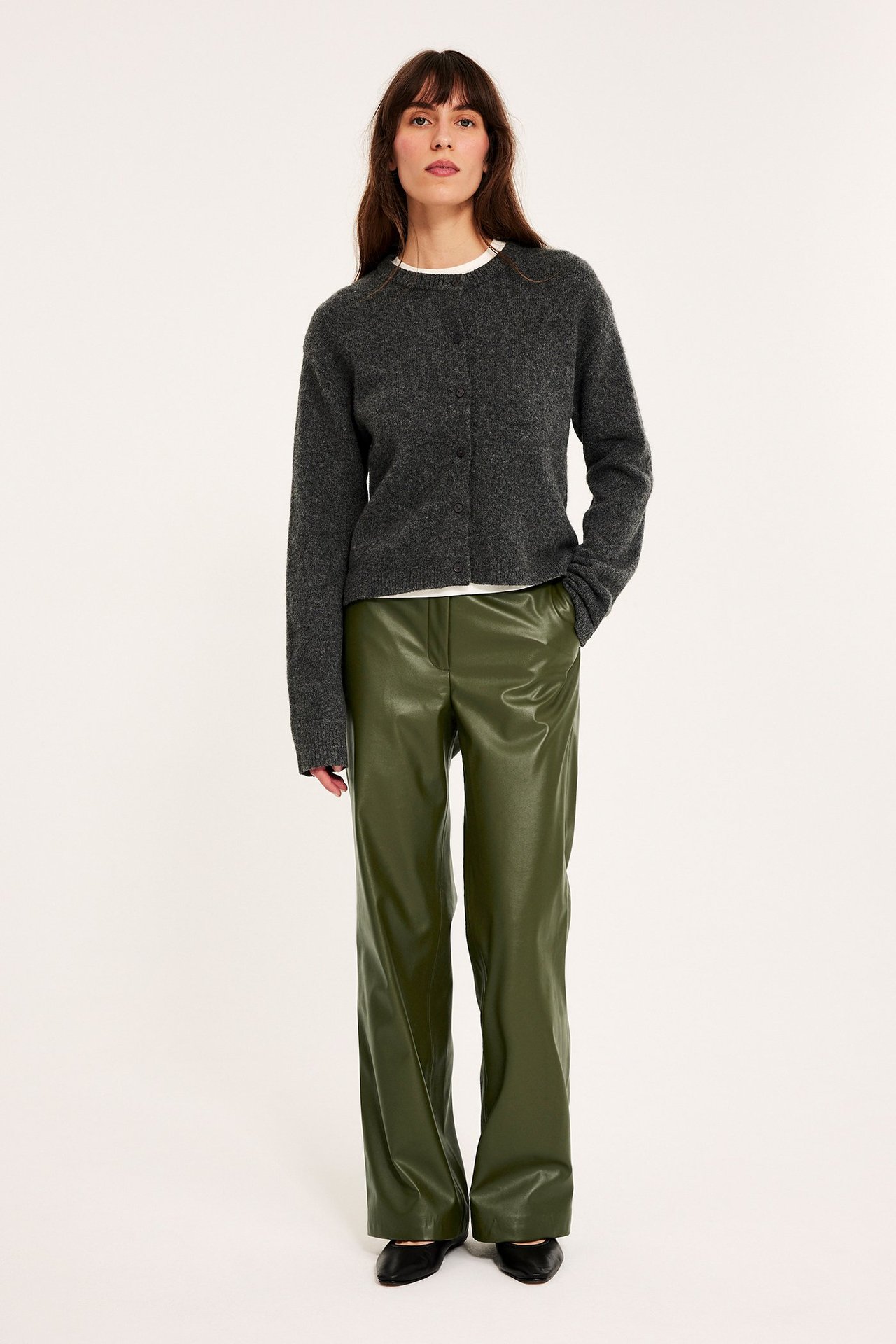 Edestäpäin: nainen yllään vihreät tekonahkaiset leveälahkeiset housut, tummanharmaa neuletakki ja valkoinen t-paita.