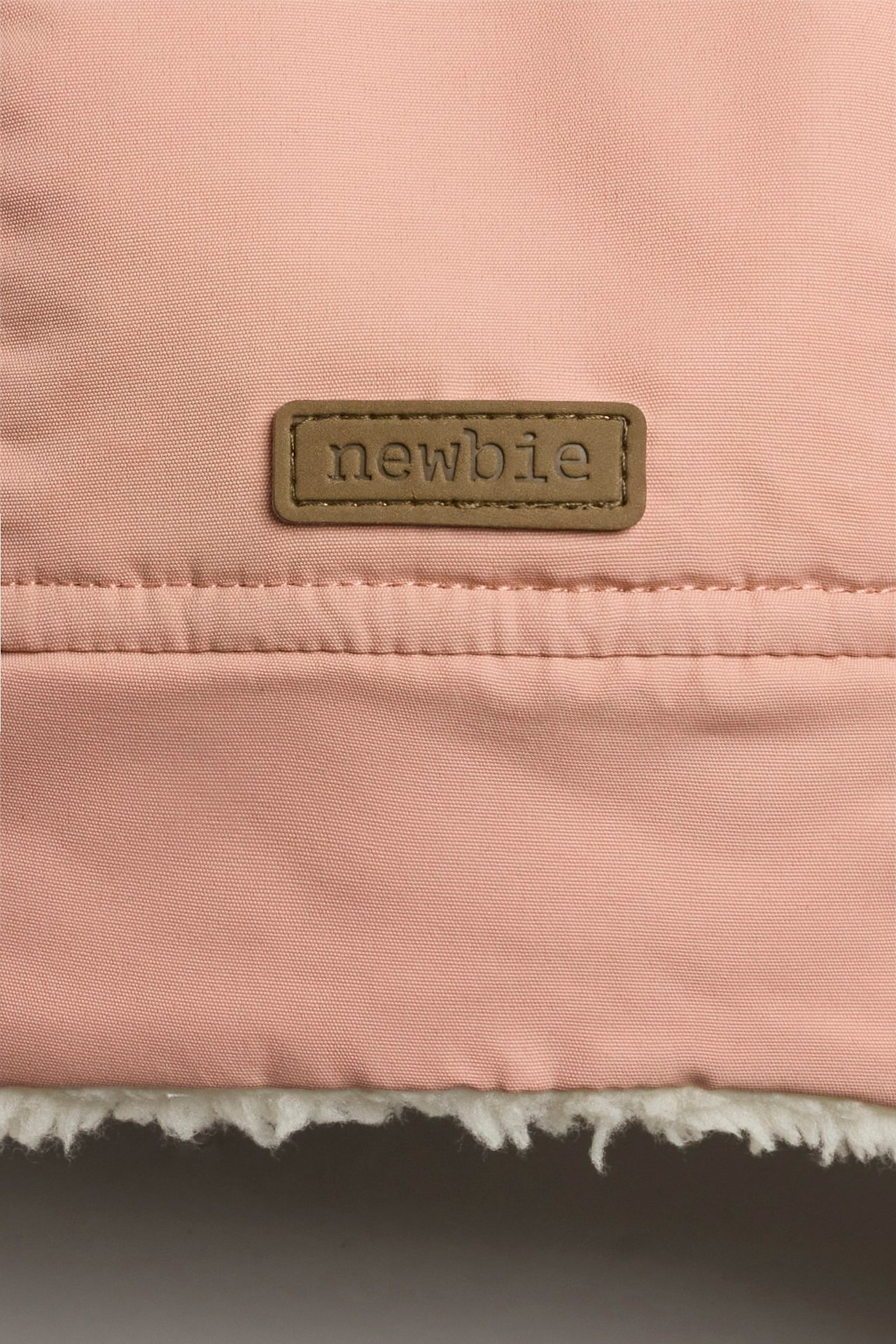 Lähikuva vaaleanpunaisen talvipipon kankaasta, jossa Newbie-merkki ja valkoinen teddyvuori.