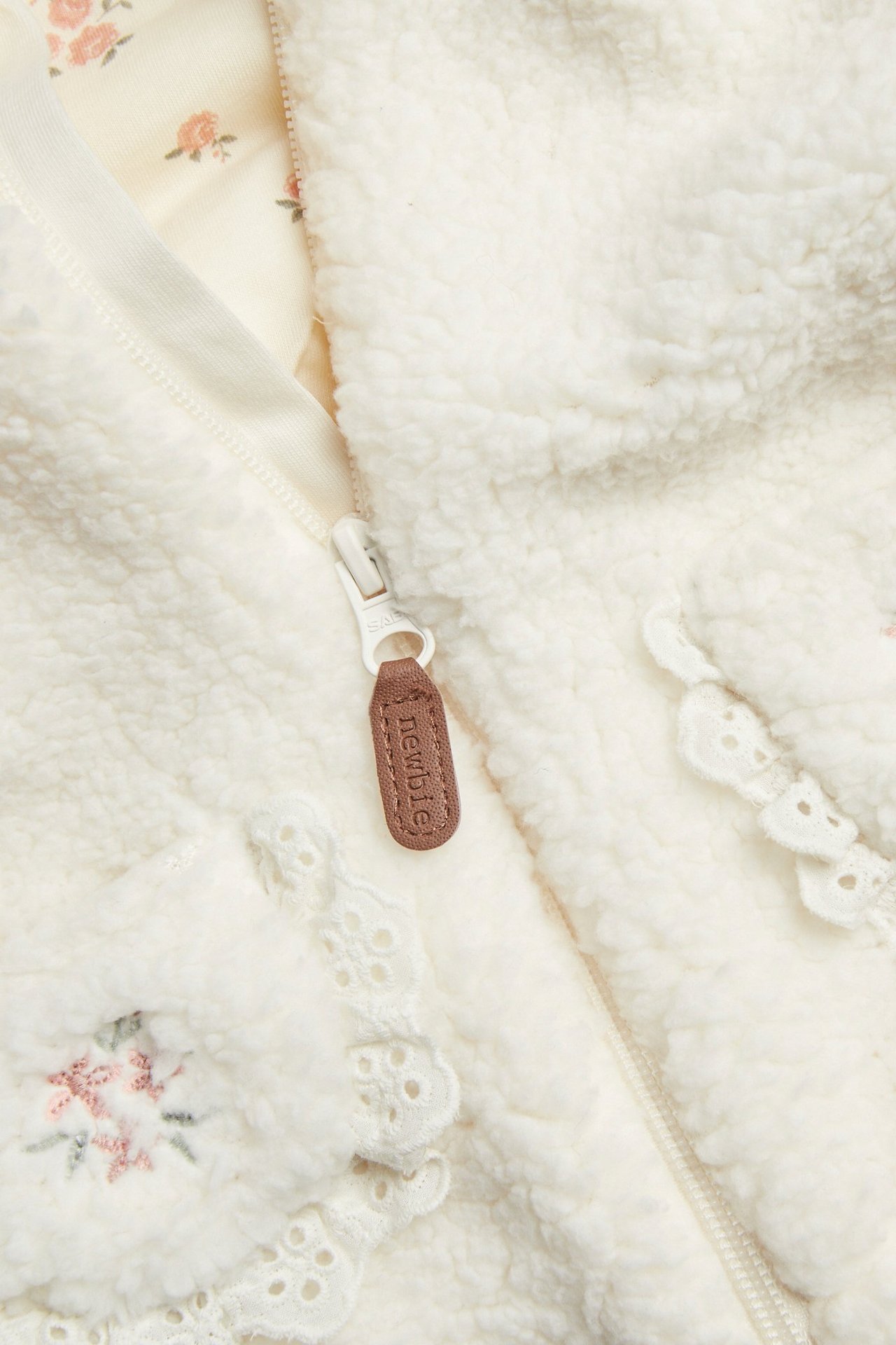 Nærbilde forfra: kremhvit barnejakke i pile med glidelås, blondekant, blomsterbroderi og mønstret fôr.