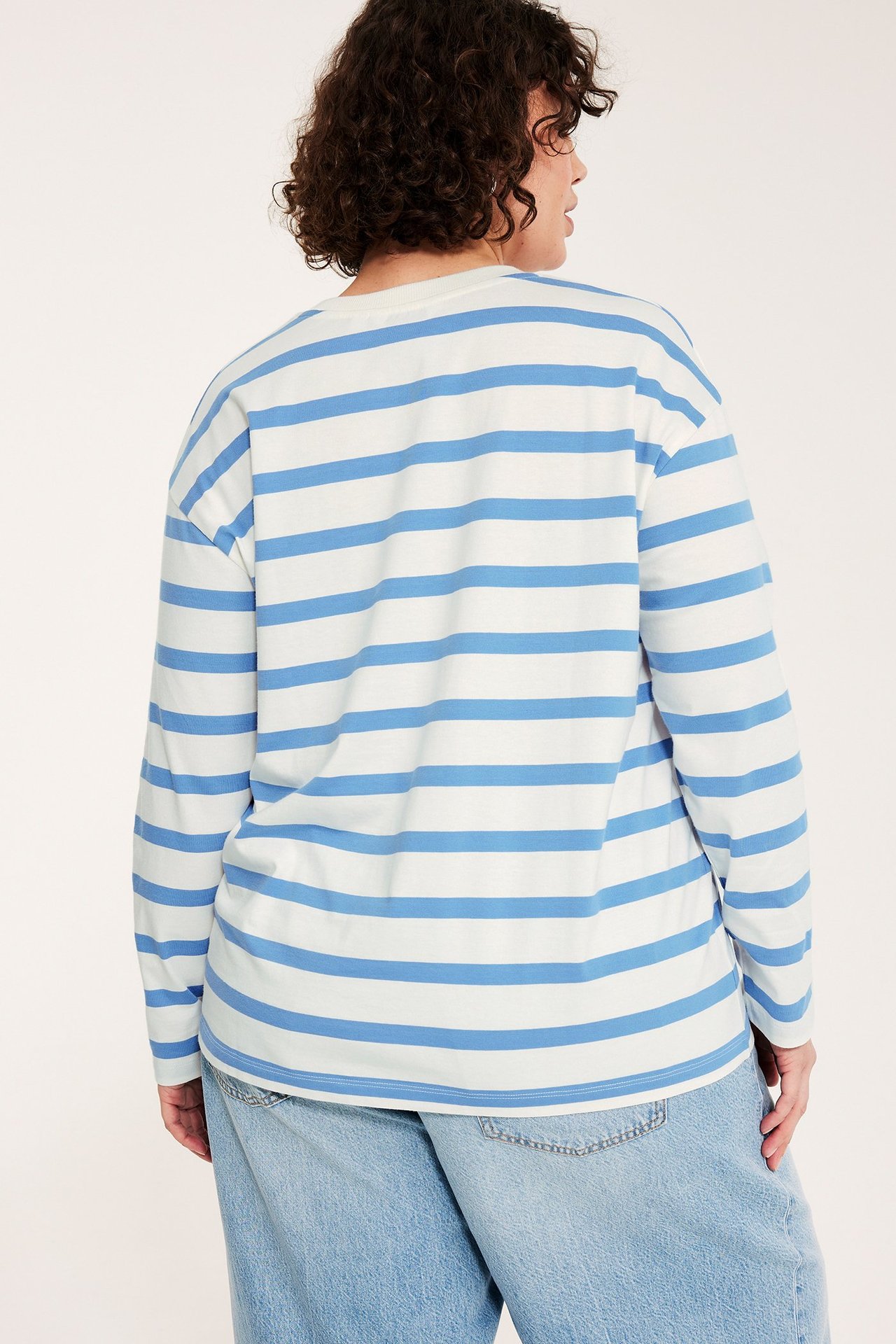 Widok z tyłu kobiety w luźnym, biało-jasnoniebieskim bawełnianym topie w paski i jasnoniebieskich jeansach.