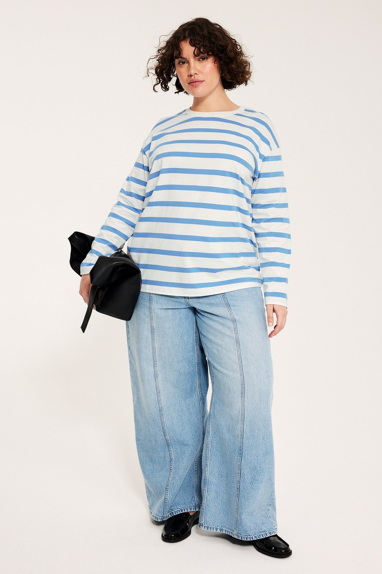 Kobieta w białym topie z długim rękawem w niebieskie paski, jasnoniebieskich szerokich jeansach, czarnych mokasynach, trzymająca czarną torebkę.