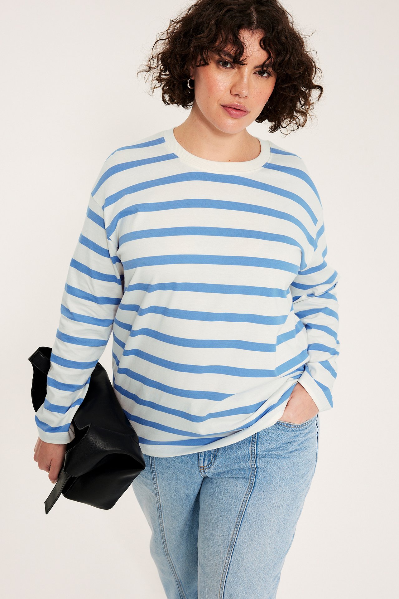 Kobieta w biało-jasnoniebieskiej bluzce w paski z długim rękawem, jasnoniebieskich jeansach, trzyma czarną torbę.