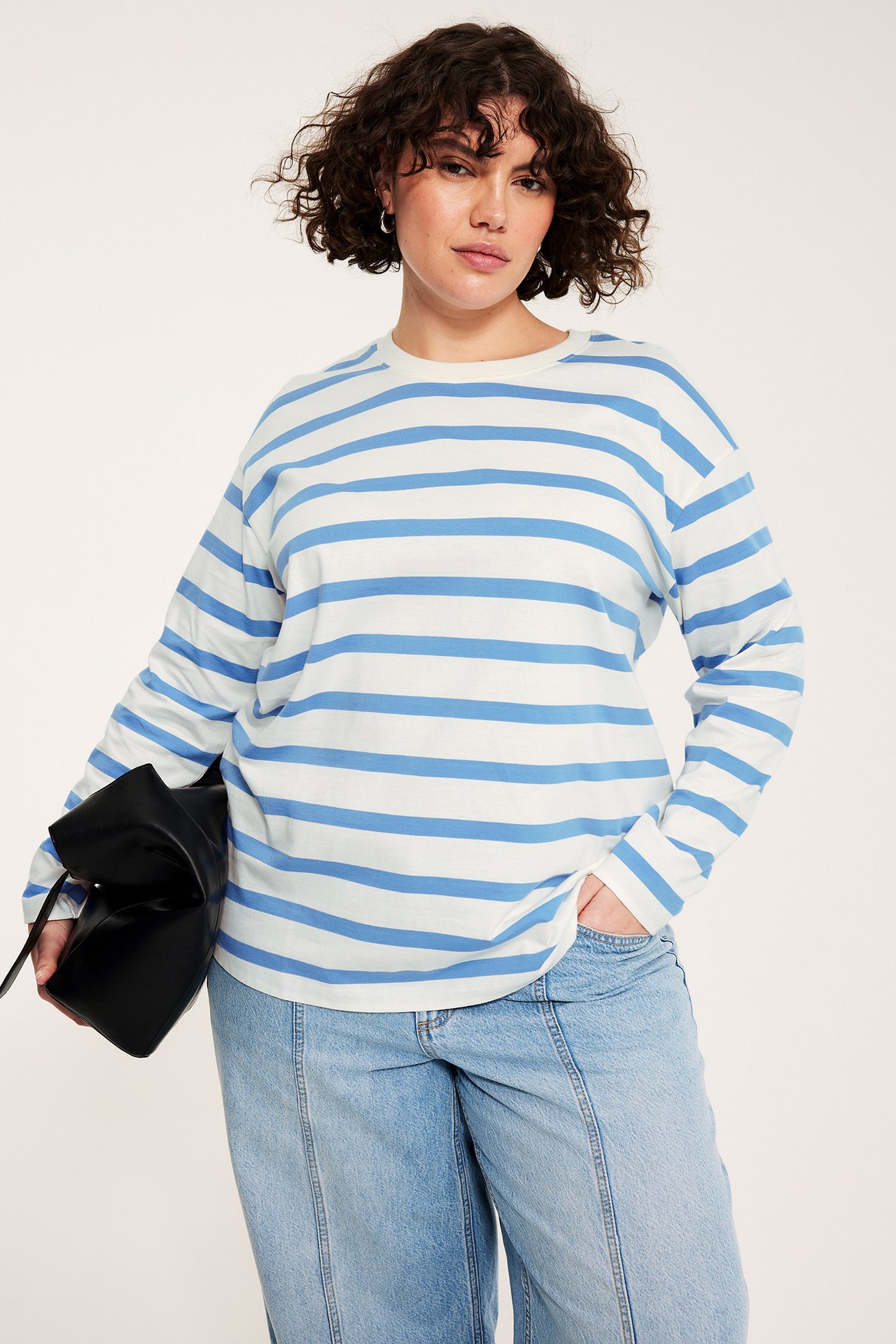Widok z przodu: Kobieta w niebiesko-białym pasiastym bawełnianym topie, jasnych dżinsach, trzyma czarną torebkę.