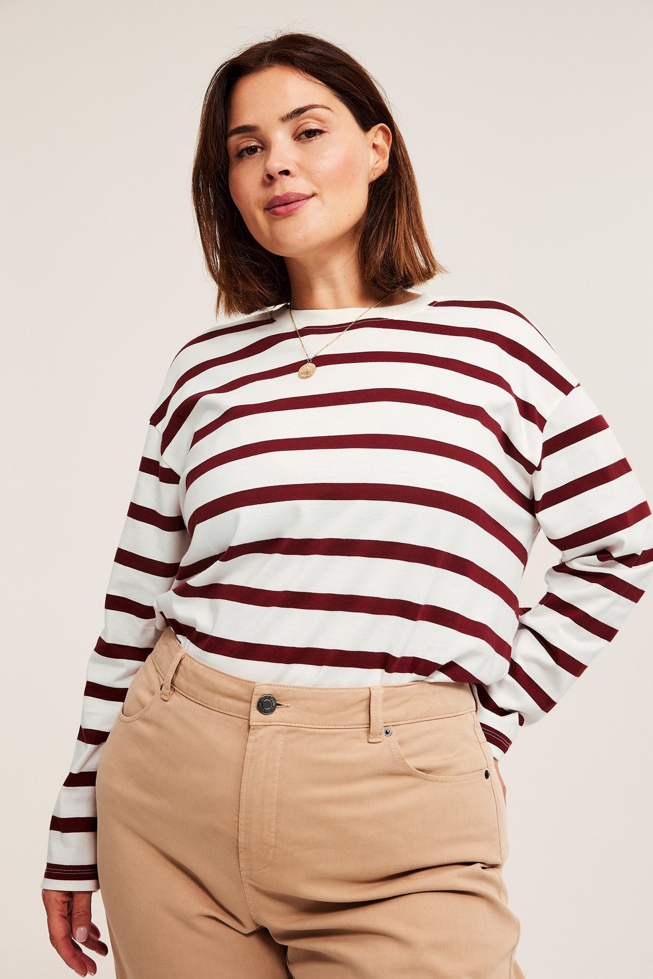 Framifrån: Kvinna i vit- och rödrandig långärmad t-shirt, beige byxor och guldhalsband.