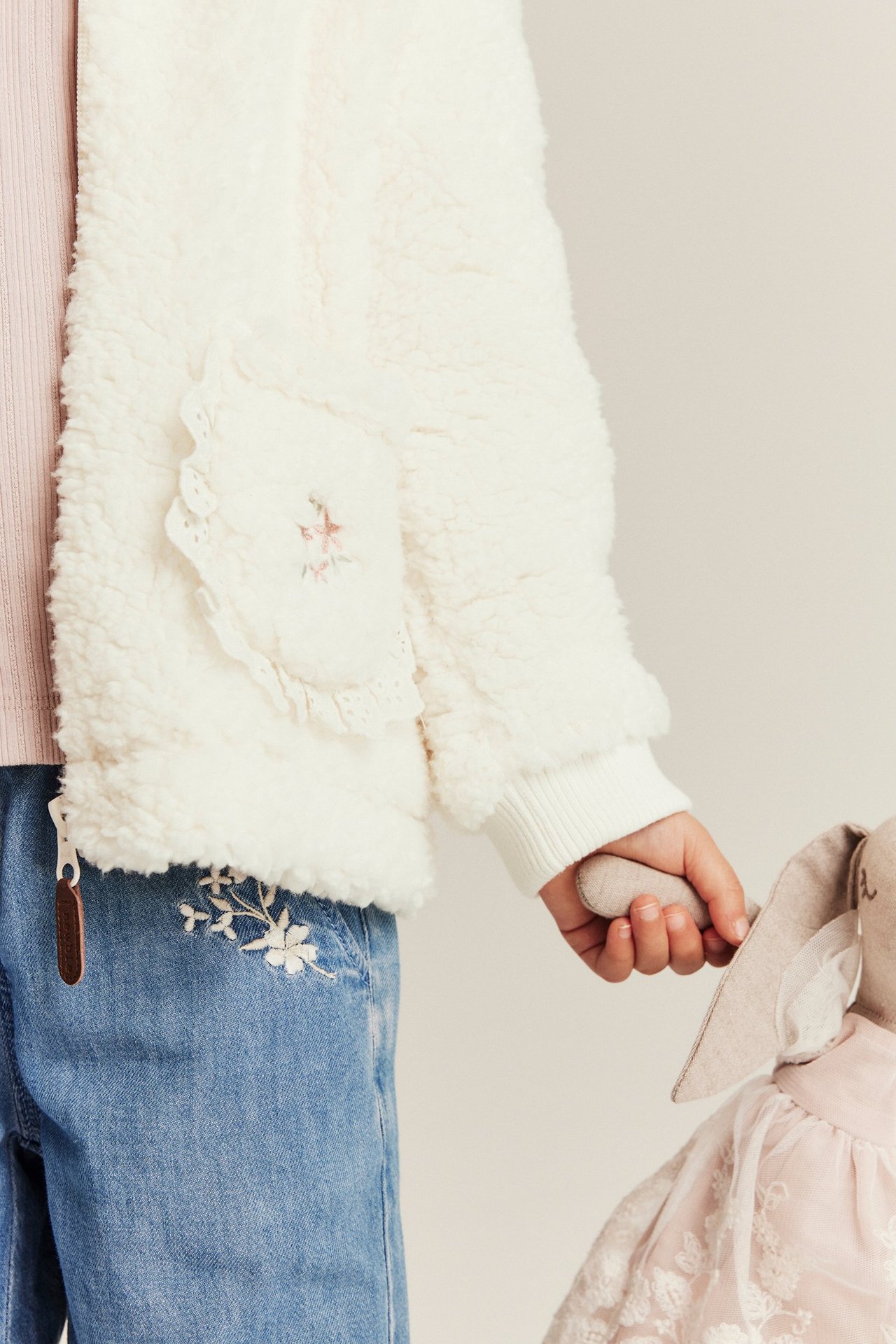 Barn iført offwhite pilejakke med blonde og broderi, rosa topp, blå broderte jeans, holder kanindukke.