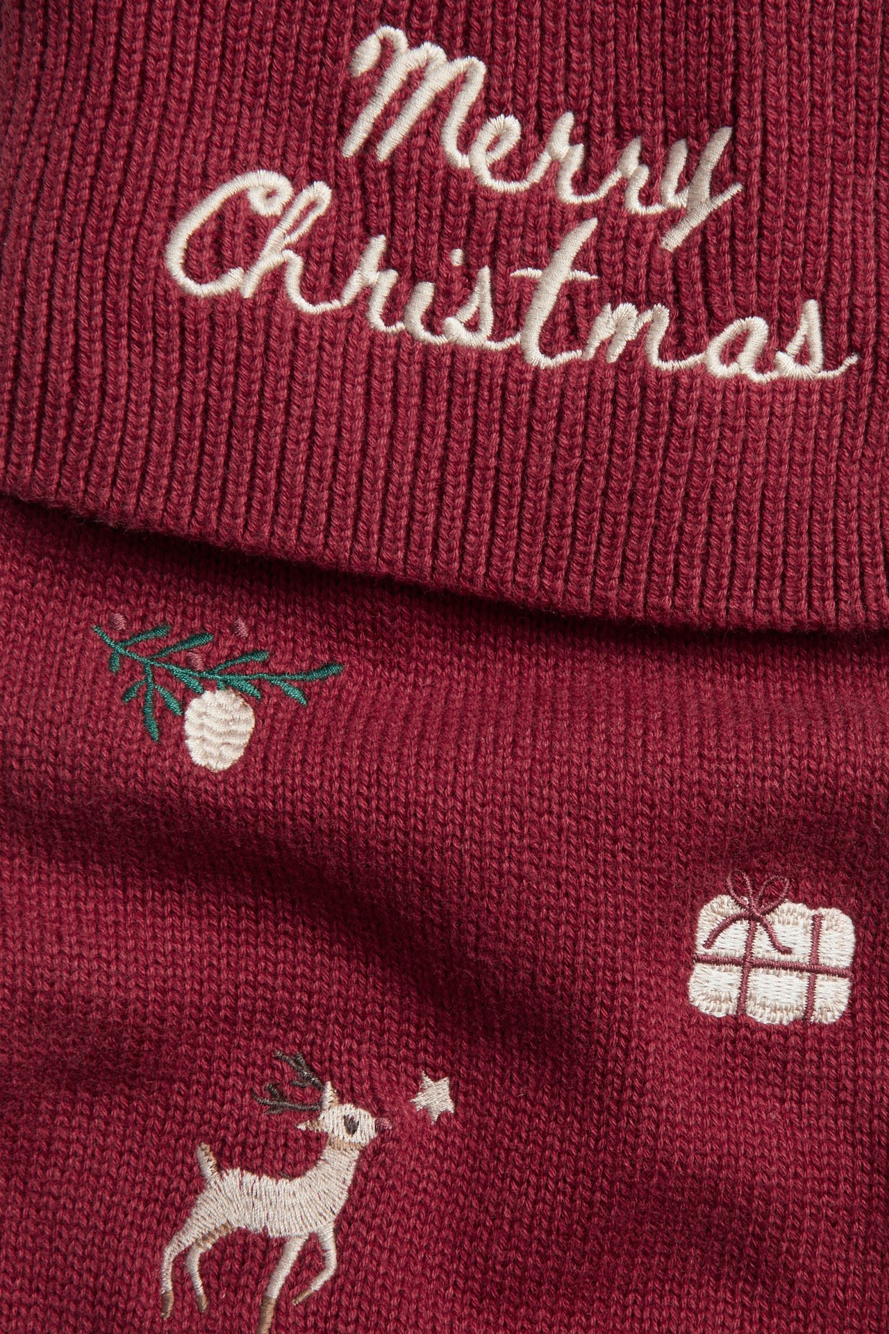 Lähikuva punaisesta neuleesta, jossa valkoinen brodeerattu 'Merry Christmas' ja juhlallisia kuvioita.