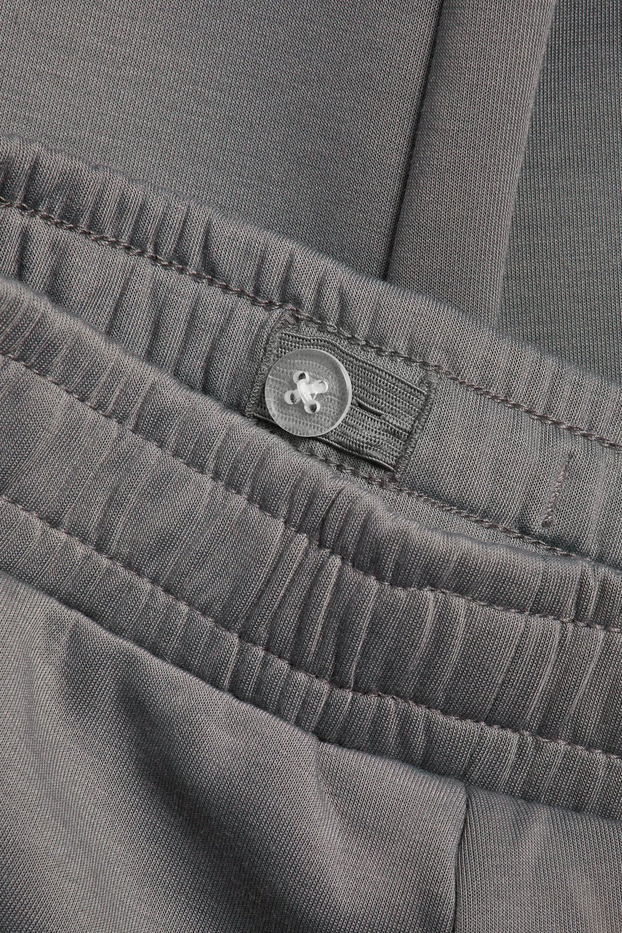 Zbliżenie szarych spodni dresowych z interlocku, widoczny elastyczny pas z przezroczystym guzikiem.