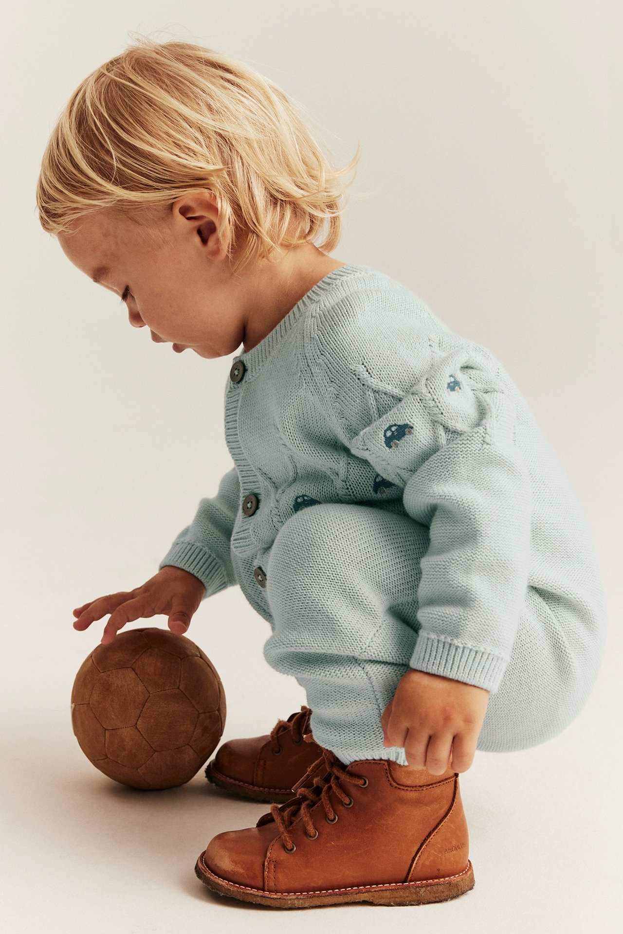 Sidebilde: Baby i lyse turkis strukturstrikket bomullsjumpsuit med bilbroderi og brune støvler.
