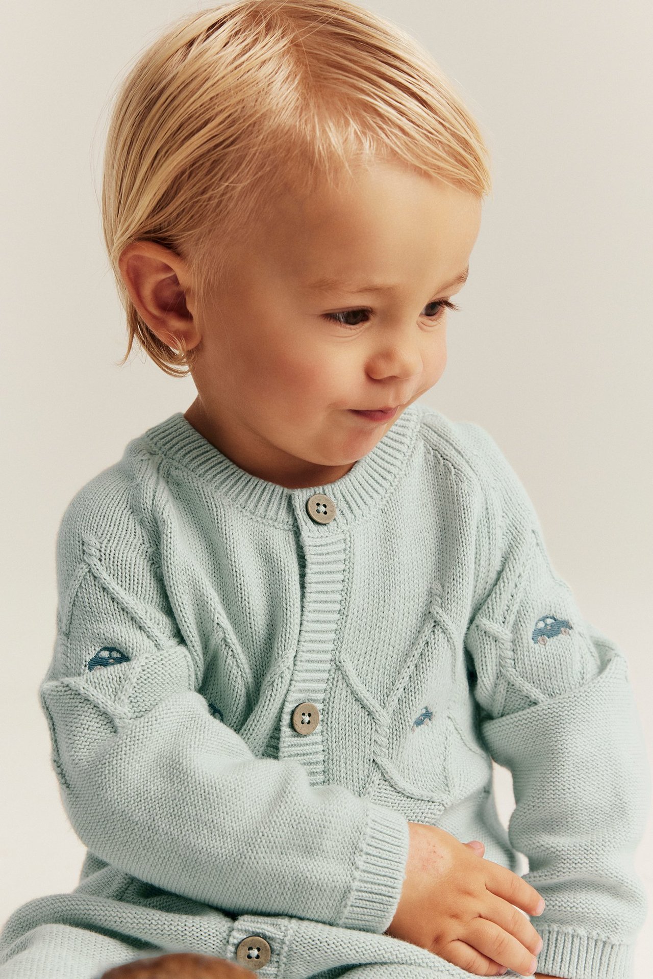 Zbliżenie dziecka w jasnoniebieskim, dzianinowym kombinezonie z haftowanymi samochodami i guzikami.
