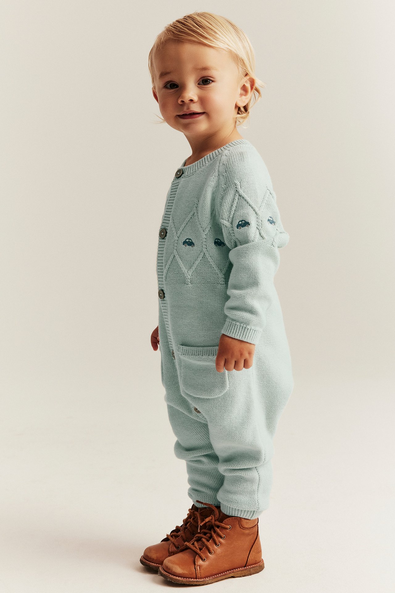 Widok z boku dziecka w jasno turkusowym, dzianinowym kombinezonie bawełnianym z haftem samochodów i brązowymi butami.