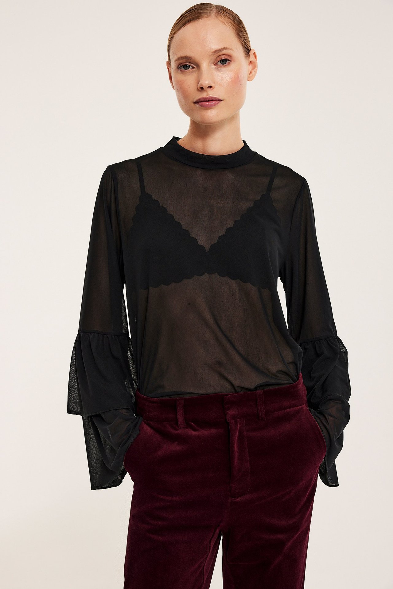 Widok z przodu, kobieta w czarnym siateczkowym topie z falbanami, czarnym biustonoszu i bordowych spodniach.