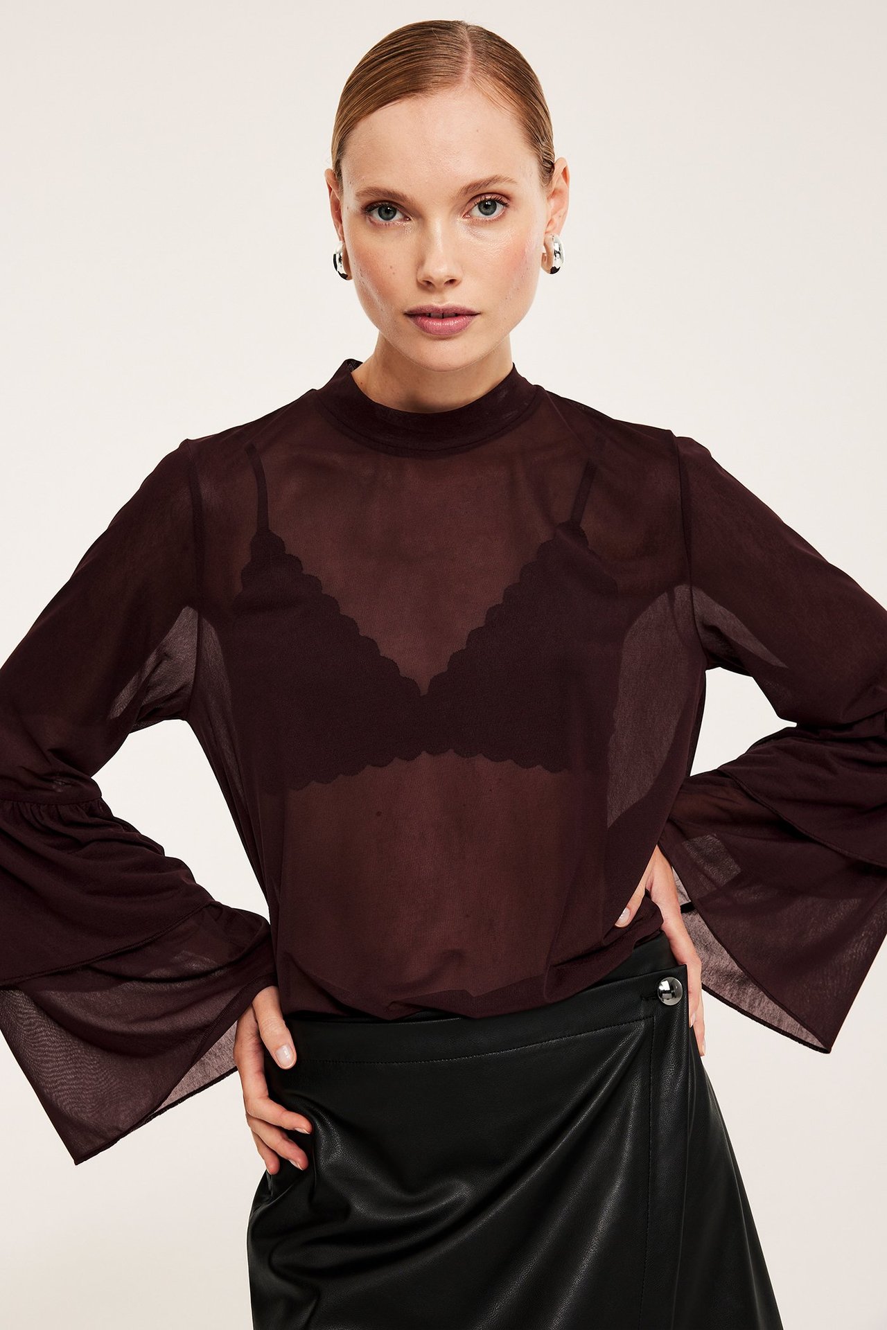 Edestä, nainen läpinäkyvässä viininpunaisessa verkkopuserossa, levenevät hihat, mustat rintaliivit, tekonahkahame, hopeakorvakorut.