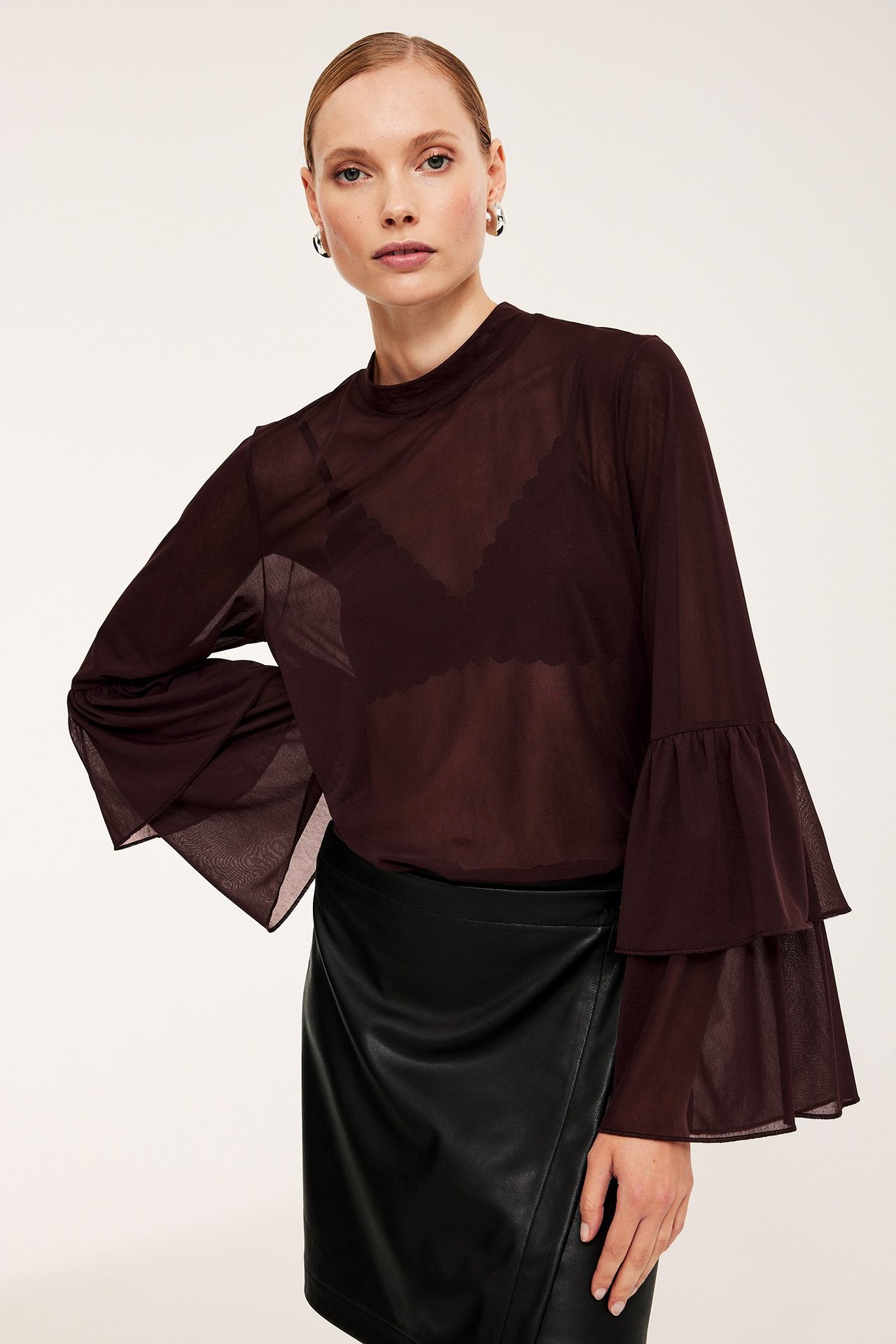 Kobieta w prześwitującej, bordowej bluzce z siateczki z falbankami, czarnym biustonoszem i czarną spódnicą ze sztucznej skóry.