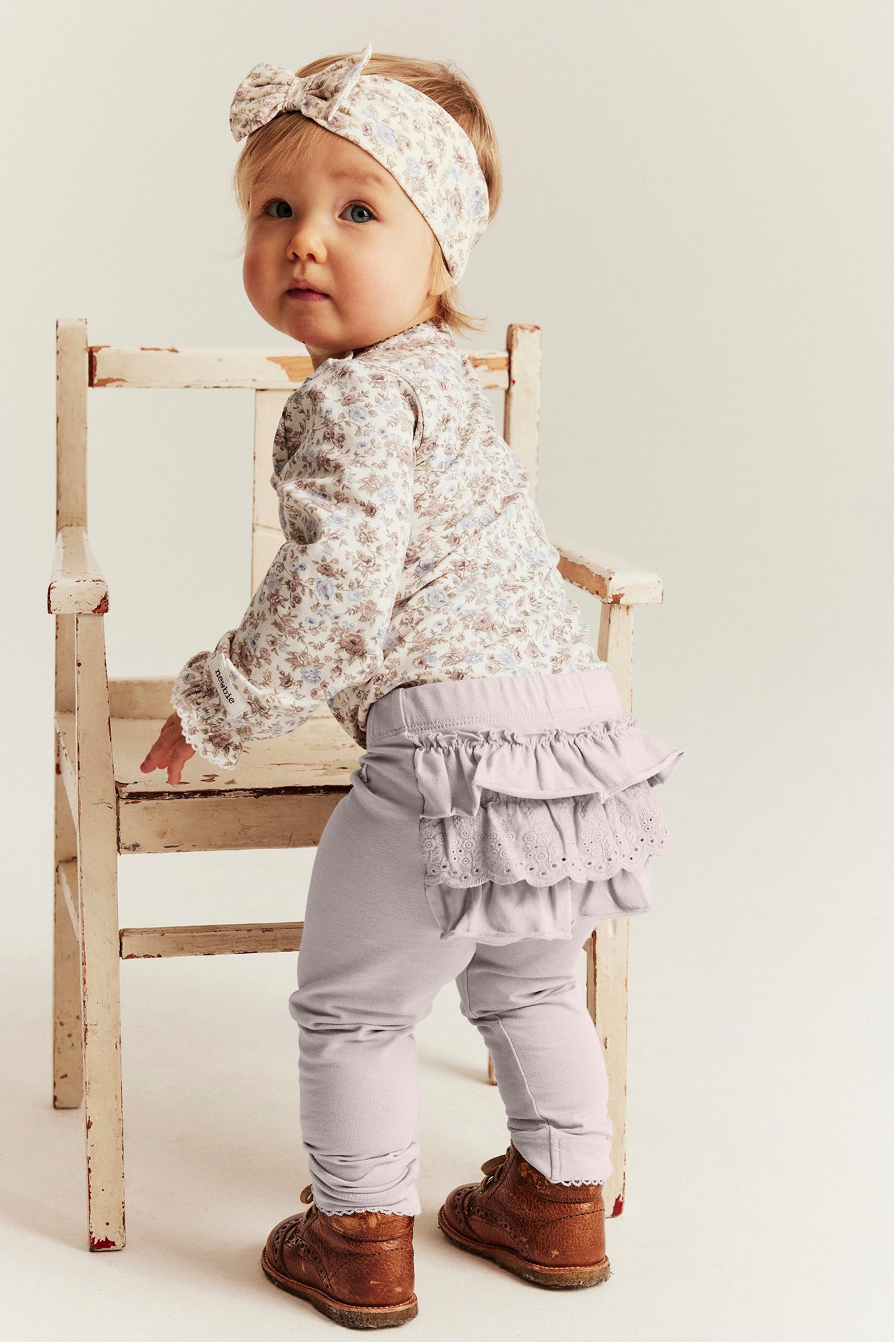 Bakfra: Baby i lys lilla volangleggings, blomstret topp, matchende hårbånd og brune sko.