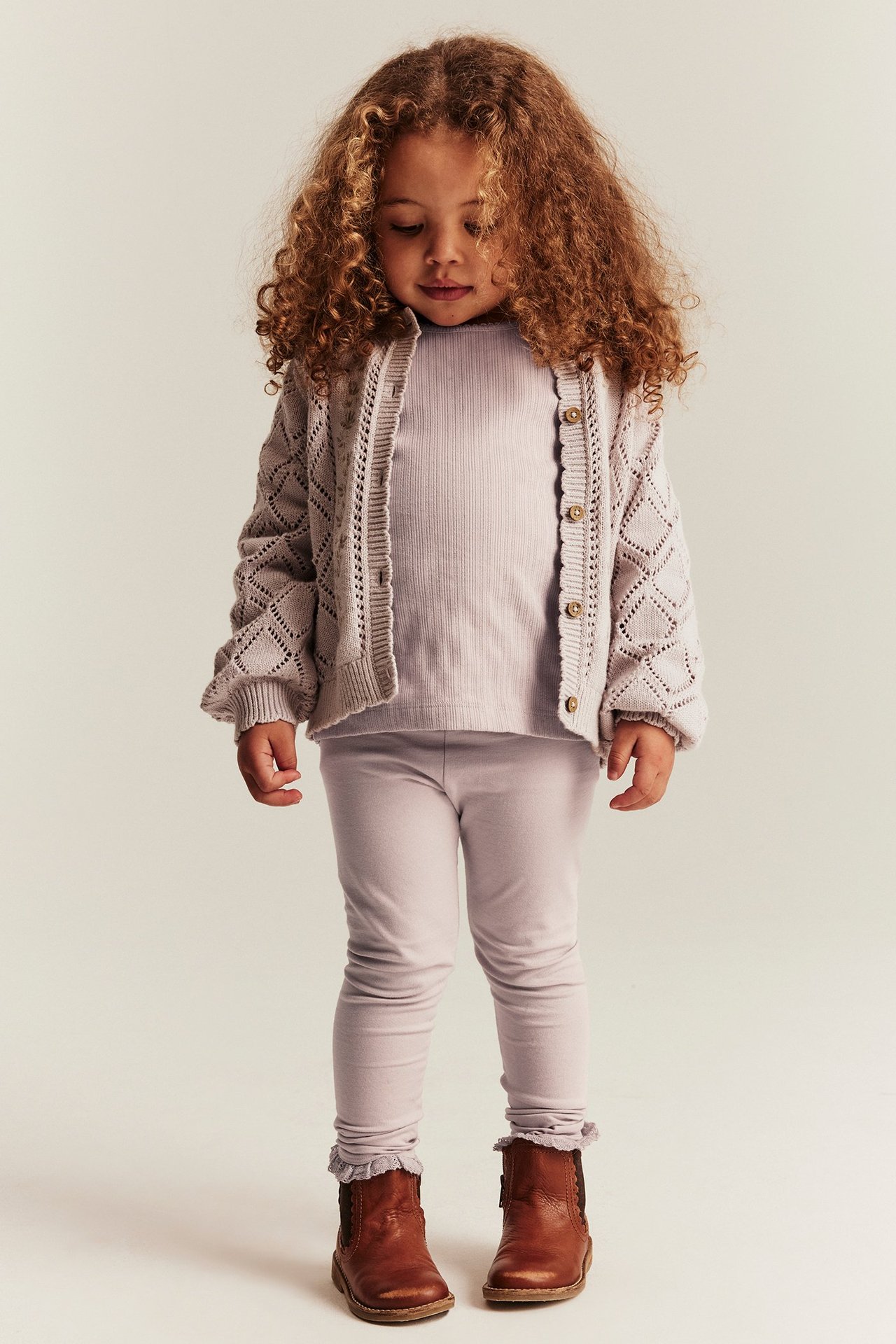 Framifrån: Barn i ljusa lila leggings med spetskant, en ribbad topp och öppen stickad kofta.