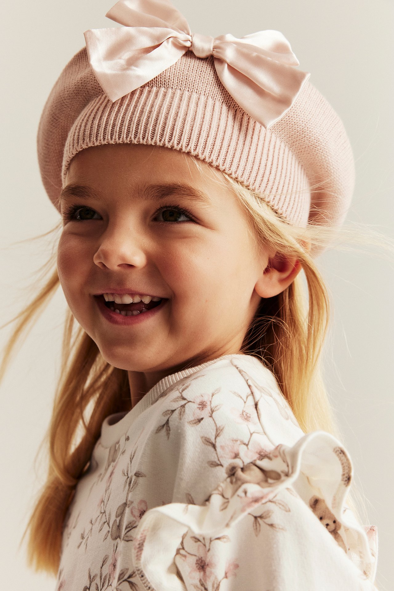 Nærbilde av en jente i offwhite genser med blomster- og bjørnemønster, volangskuldre, og rosa strikket beret.