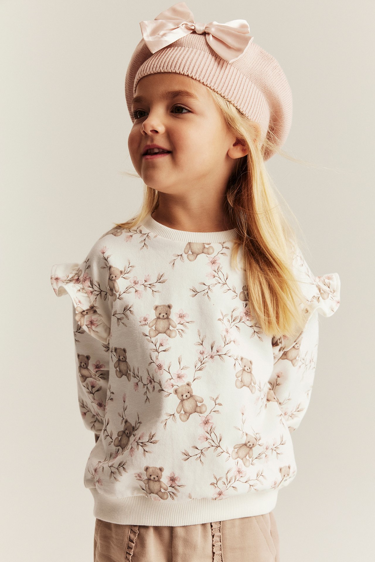 Widok z przodu: Dziecko w kremowej bawełnianej bluzie z misiami, jasnobrązowych spodniach i różowym berecie.