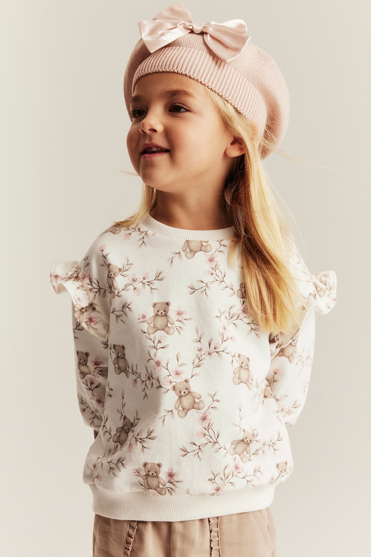 Widok z przodu: Dziecko w kremowej bawełnianej bluzie z misiami, jasnobrązowych spodniach i różowym berecie.