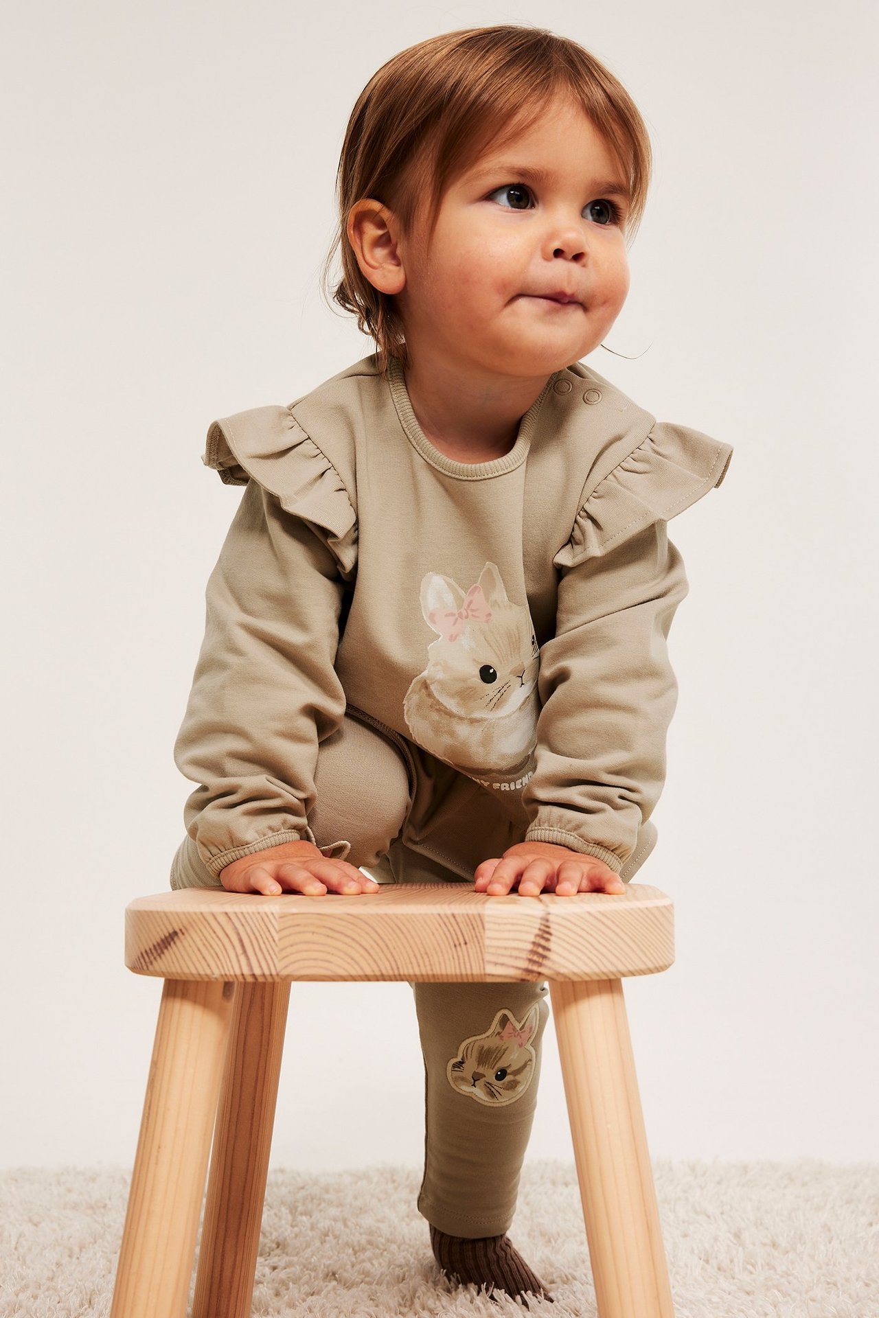 Framifrån, baby i beige volangtröja med kanintryck, matchande byxor, står på en pall.