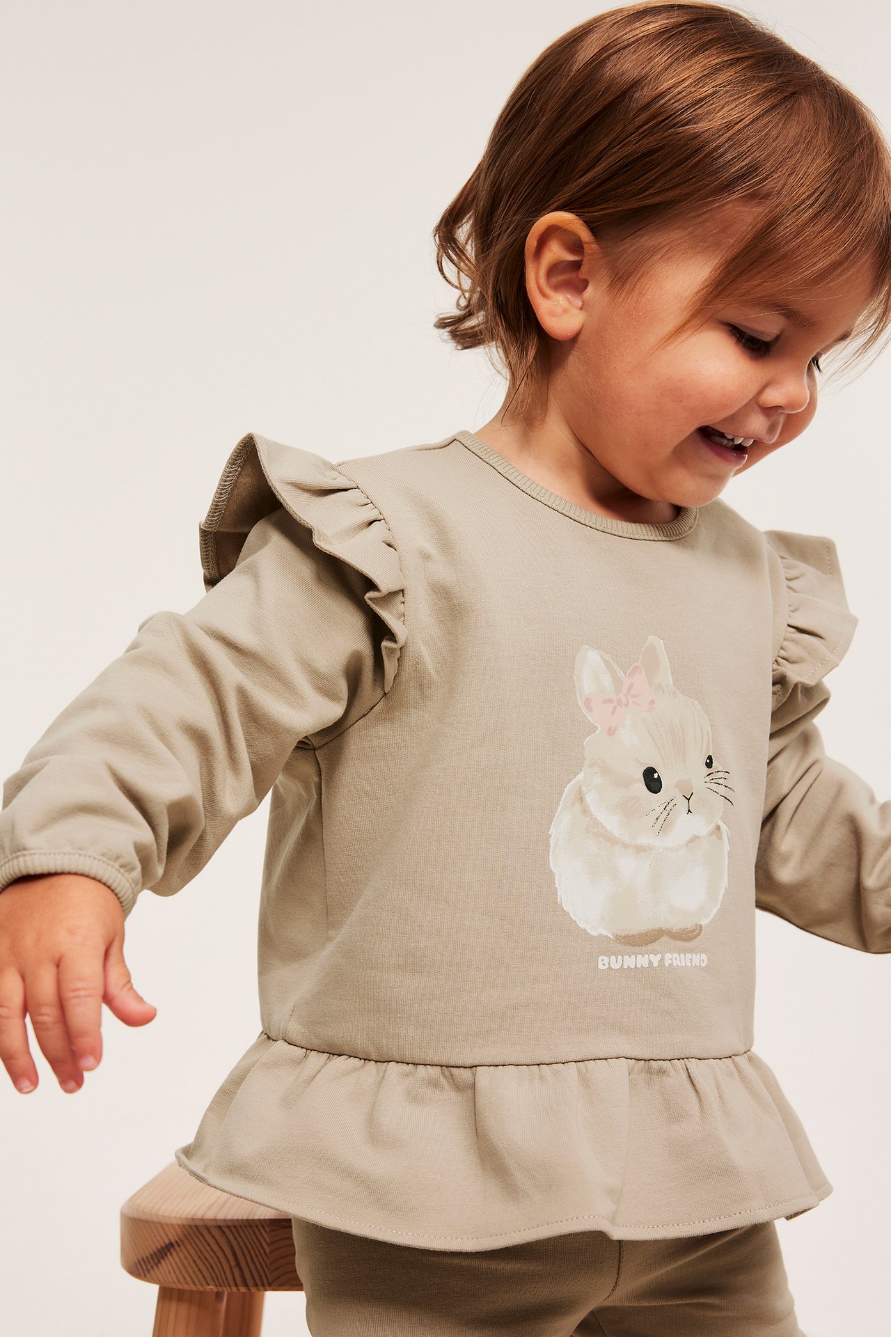 Framsidesvy av ett barn i beige sweatshirt med volanger på axlarna, kanintryck och volang nedtill.