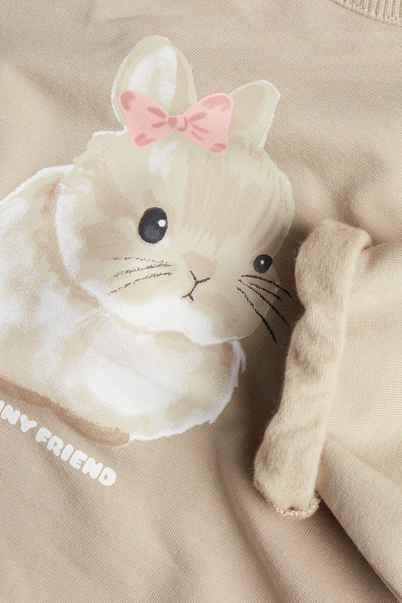 Närbild framifrån på en beige babysweatshirt med tryckt kanin, rosa rosett och beige volang.