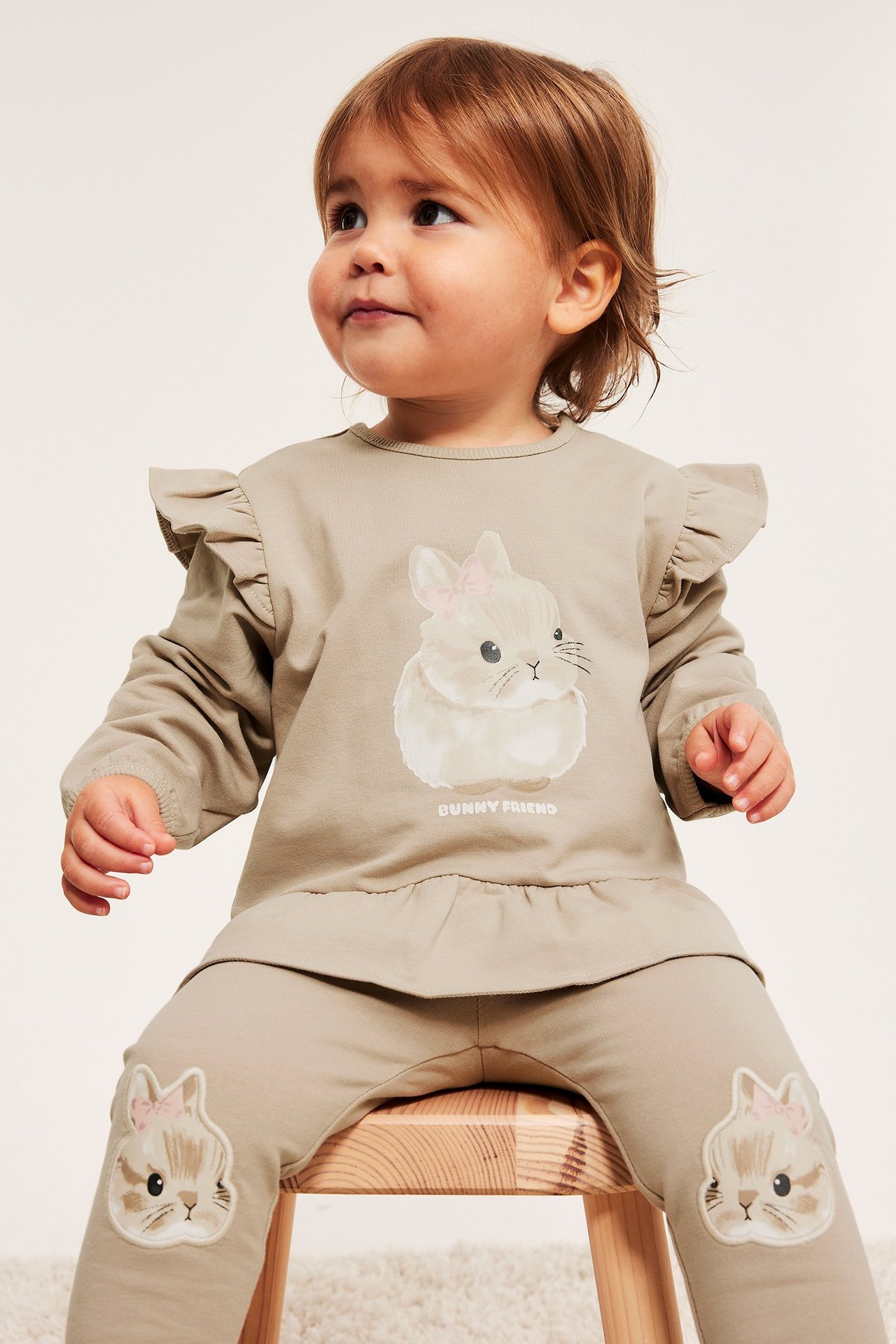 Framifrån, baby i beige bomullsblandad sweatshirt med volanger, kanintryck och matchande leggings.