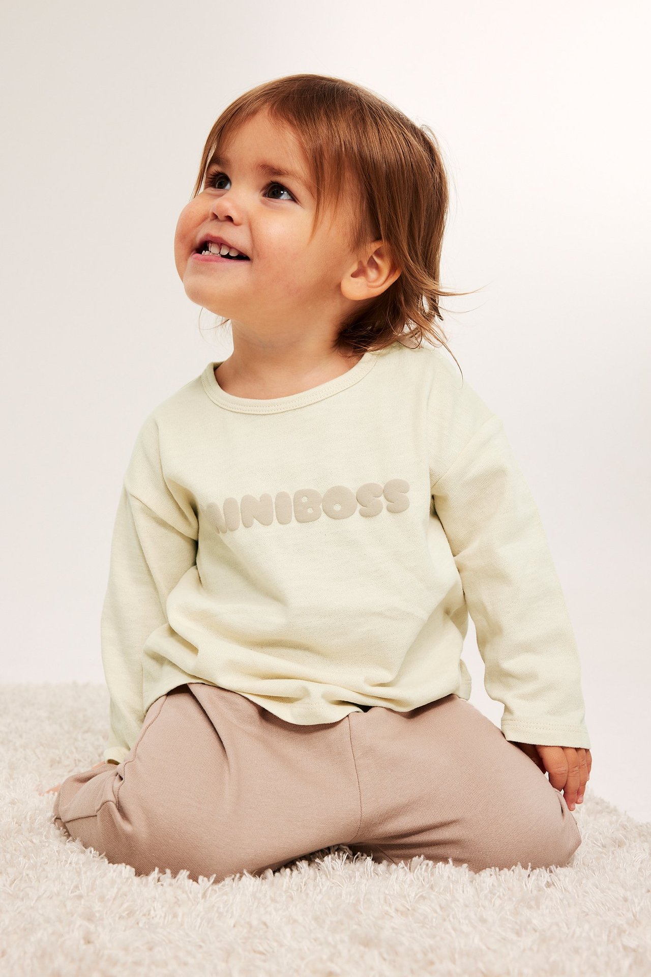 Framifrån, baby i ljusbeige bomulls långärmad t-shirt med MINIBOSS-tryck och ljusbruna byxor.