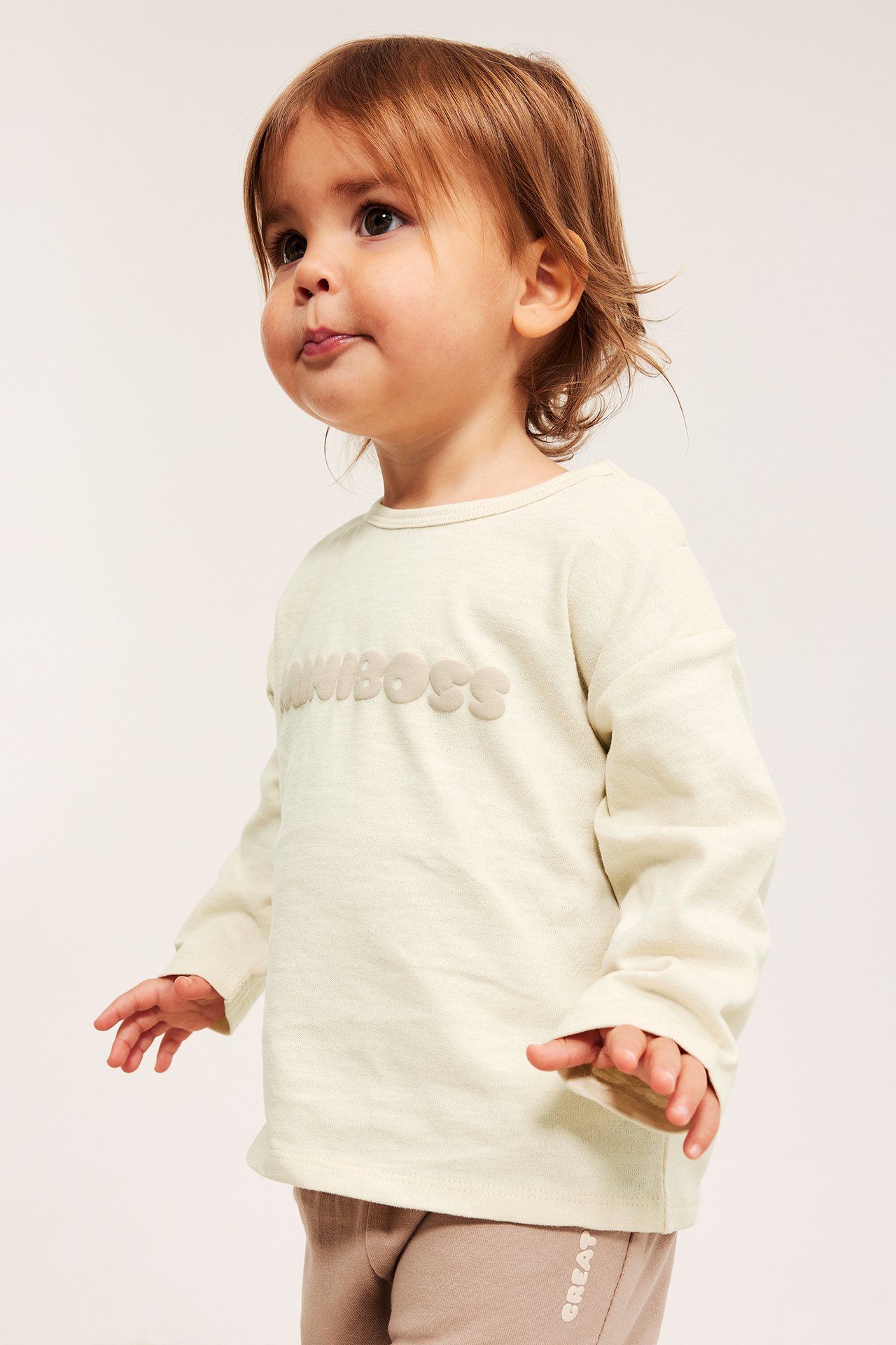 Framifrån på baby i ljusbeige bomulls t-shirt med lång ärm och 'MINIBOSS'-tryck, samt ljusbruna byxor.