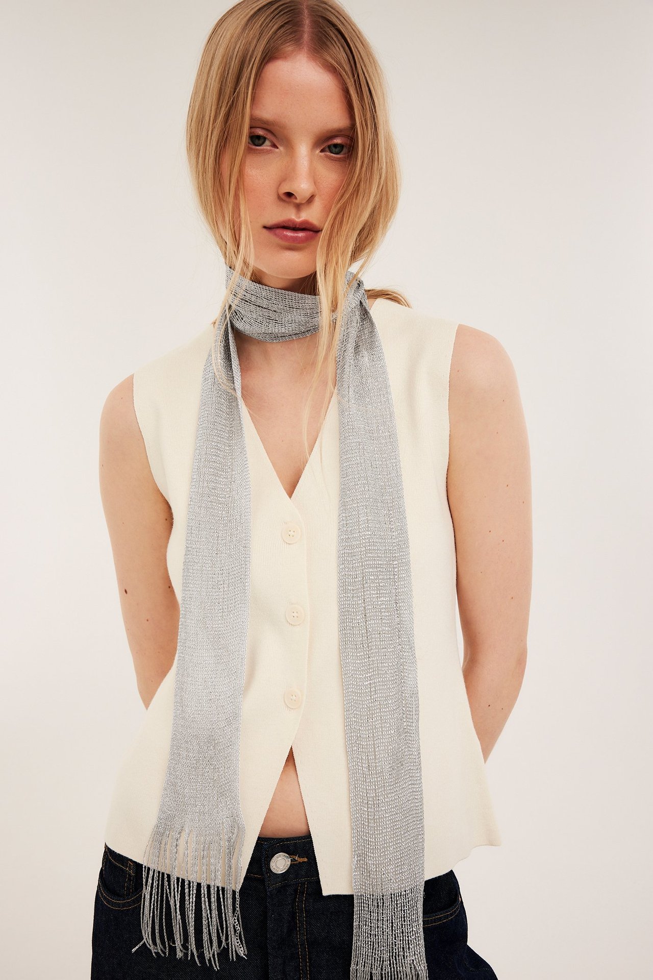 Naisen etunäkymä, hopeinen kimaltava hapsuhuivi, hihaton kermanvärinen toppi ja tummat farkut.