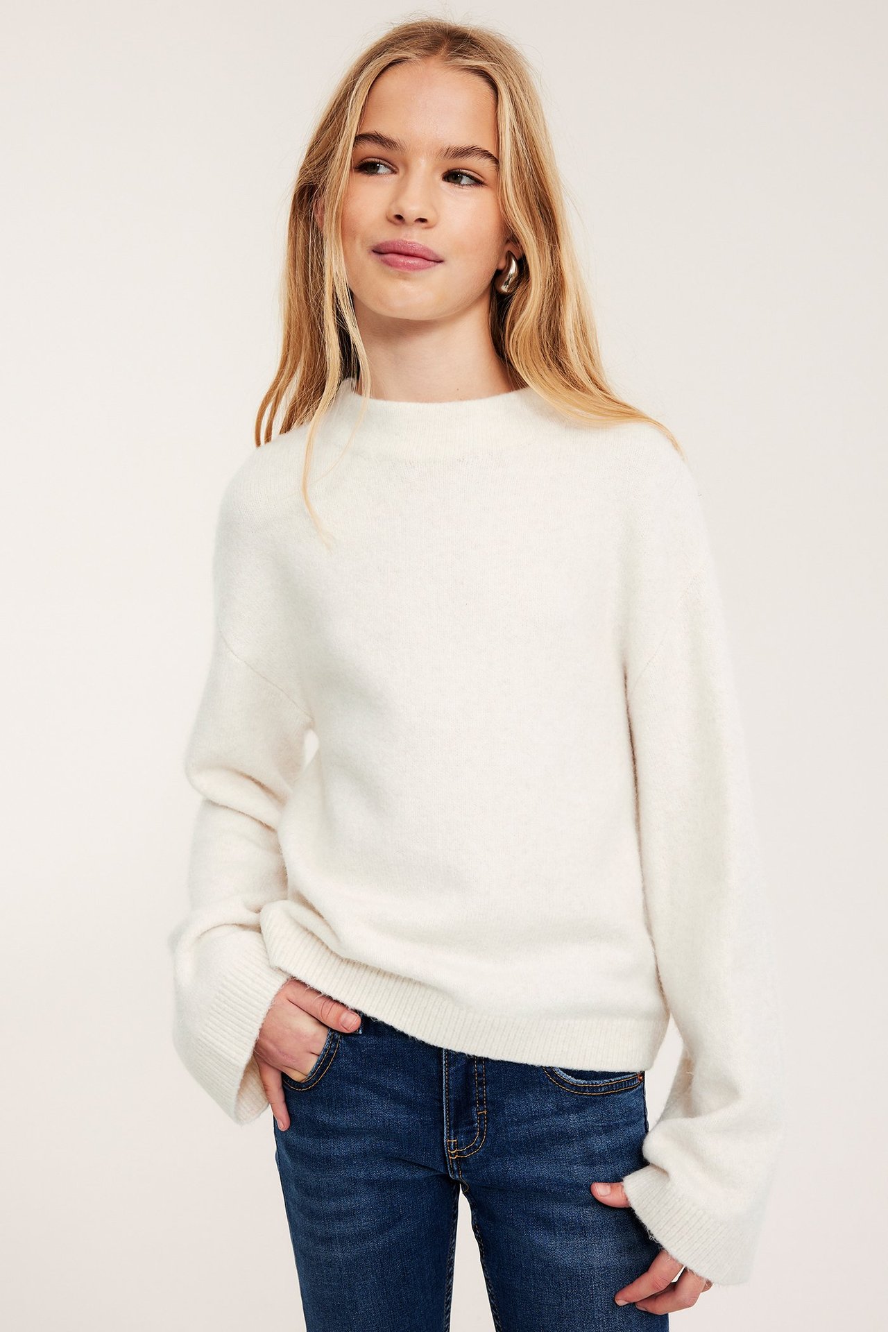 Edestä: lapsi käyttää valkoista korkeakaulaista neuletta, tummansinisiä farkkuja ja kultaisia korvakoruja.