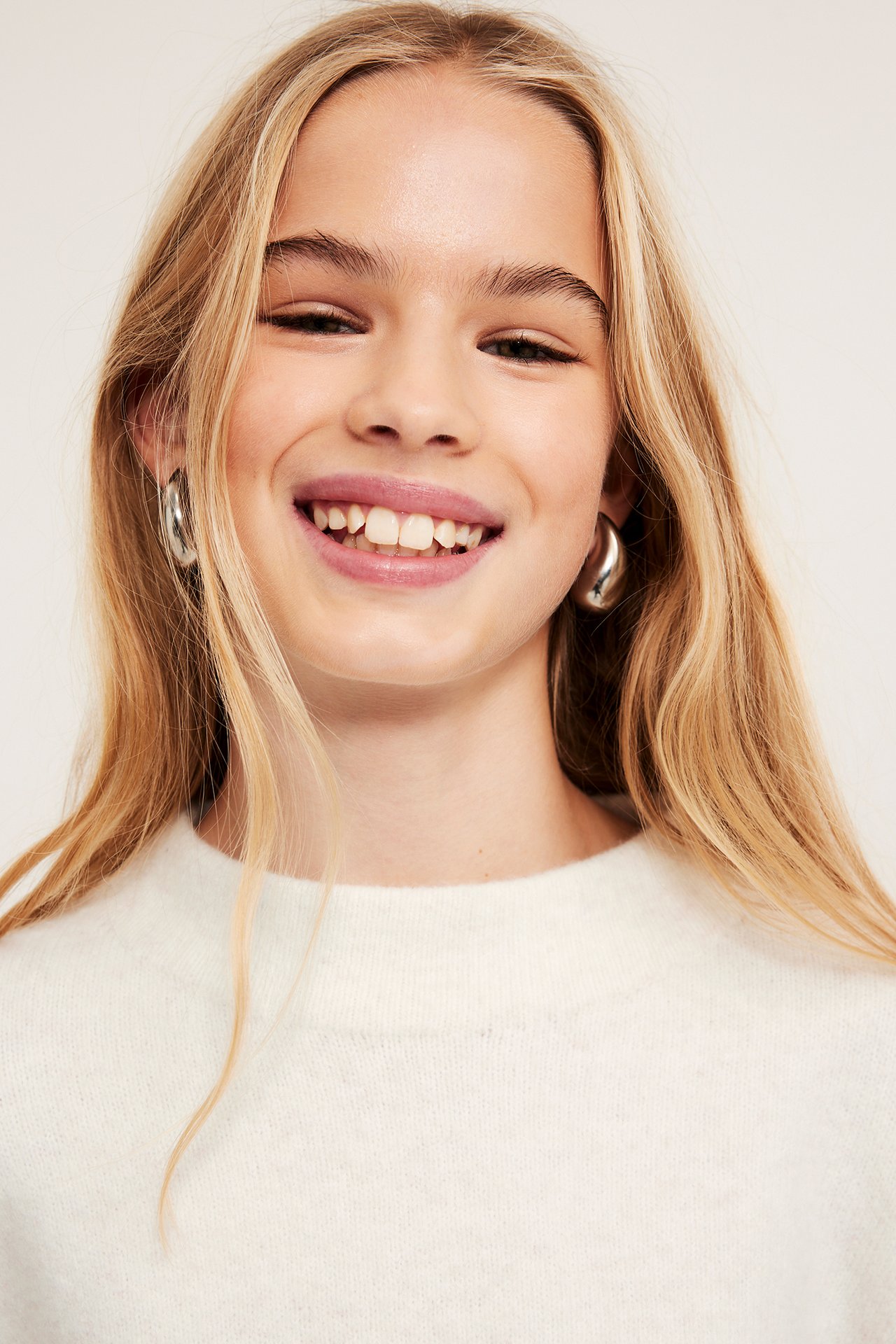 Zbliżenie z przodu dziecka w białym melanżowym swetrze dzianinowym z kominowym kołnierzem i srebrnych kolczykach kołach.