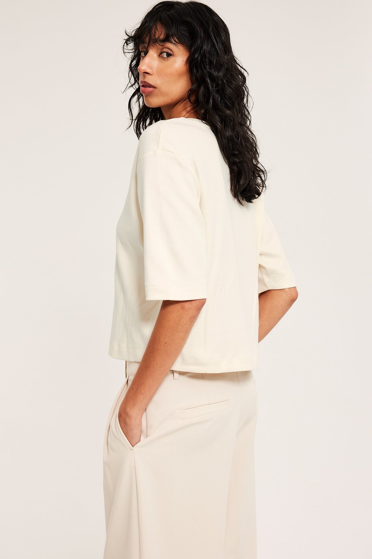 Sidovy av kvinna i off-white, enfärgad, croppad t-shirt i bomull och ljusbeige vida byxor.