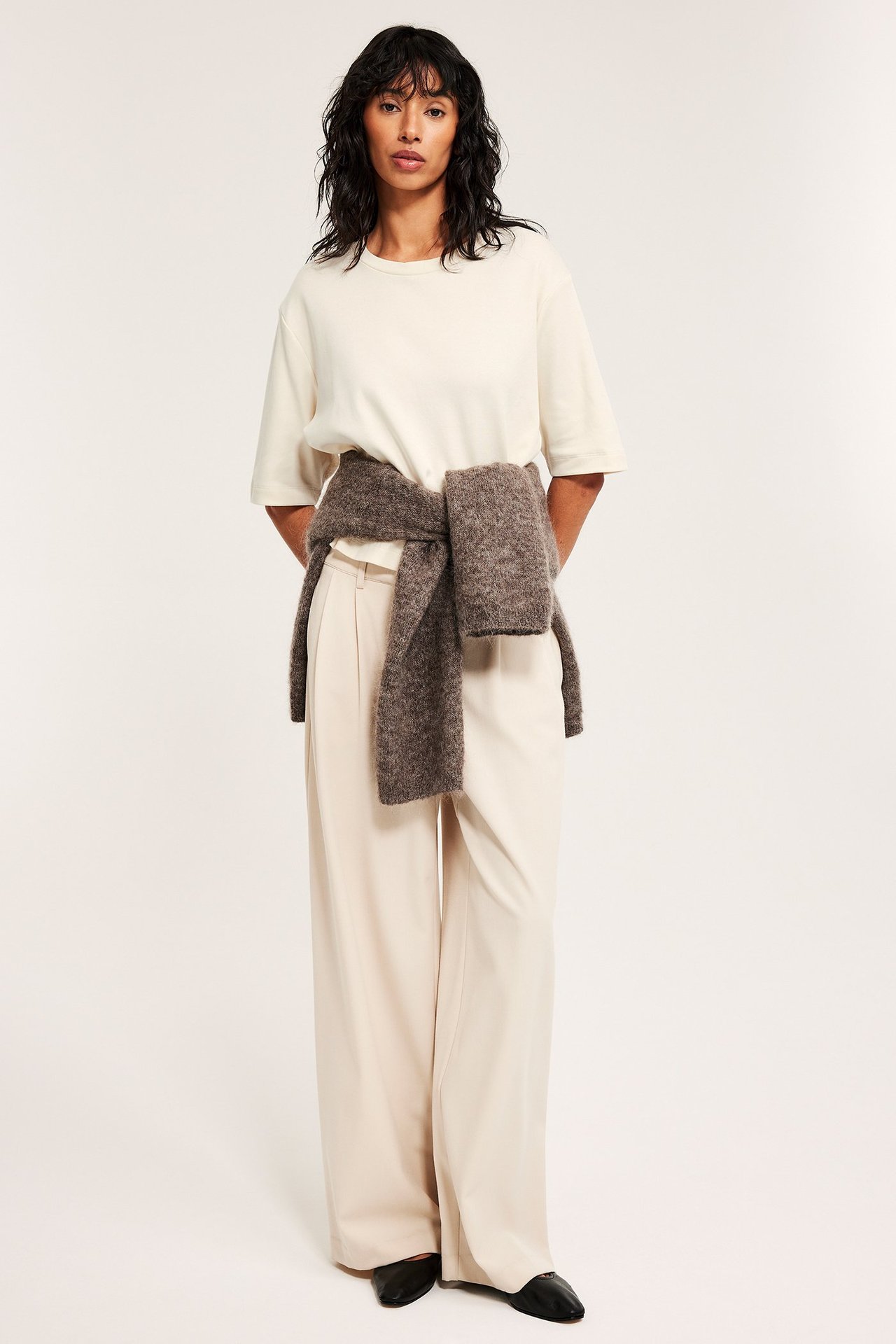 Kvinna i offwhite croppad bomulls-t-shirt, ljusbeige vida byxor, mörkbrun tröja knuten runt midjan.