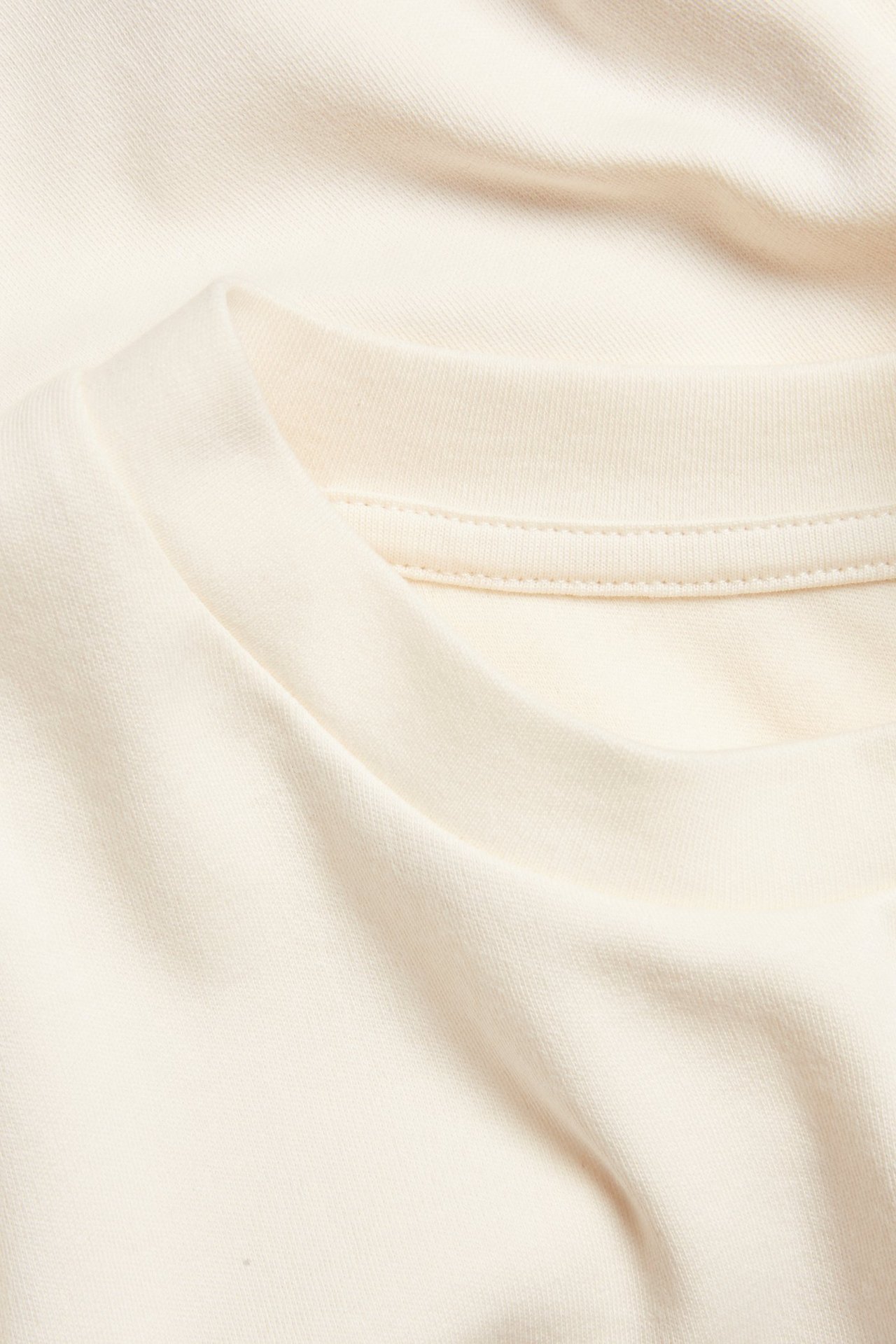 Zbliżenie na okrągły dekolt bawełnianego T-shirtu w kolorze ecru, ukazujące fakturę materiału i szwy.