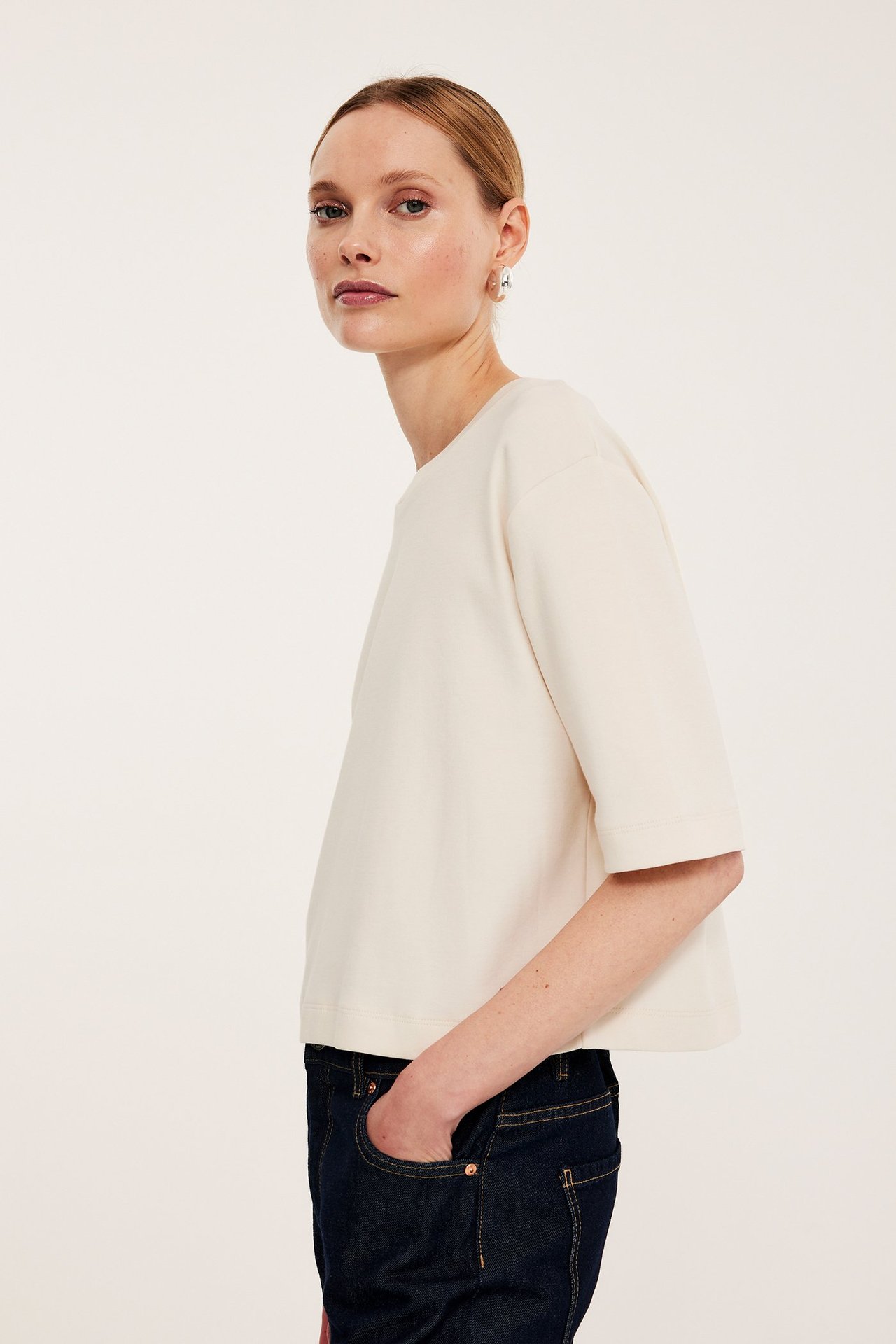 Widok z boku kobiety w kremowym, krótkim bawełnianym T-shircie, ciemnoniebieskich dżinsach i małych kolczykach kołach.