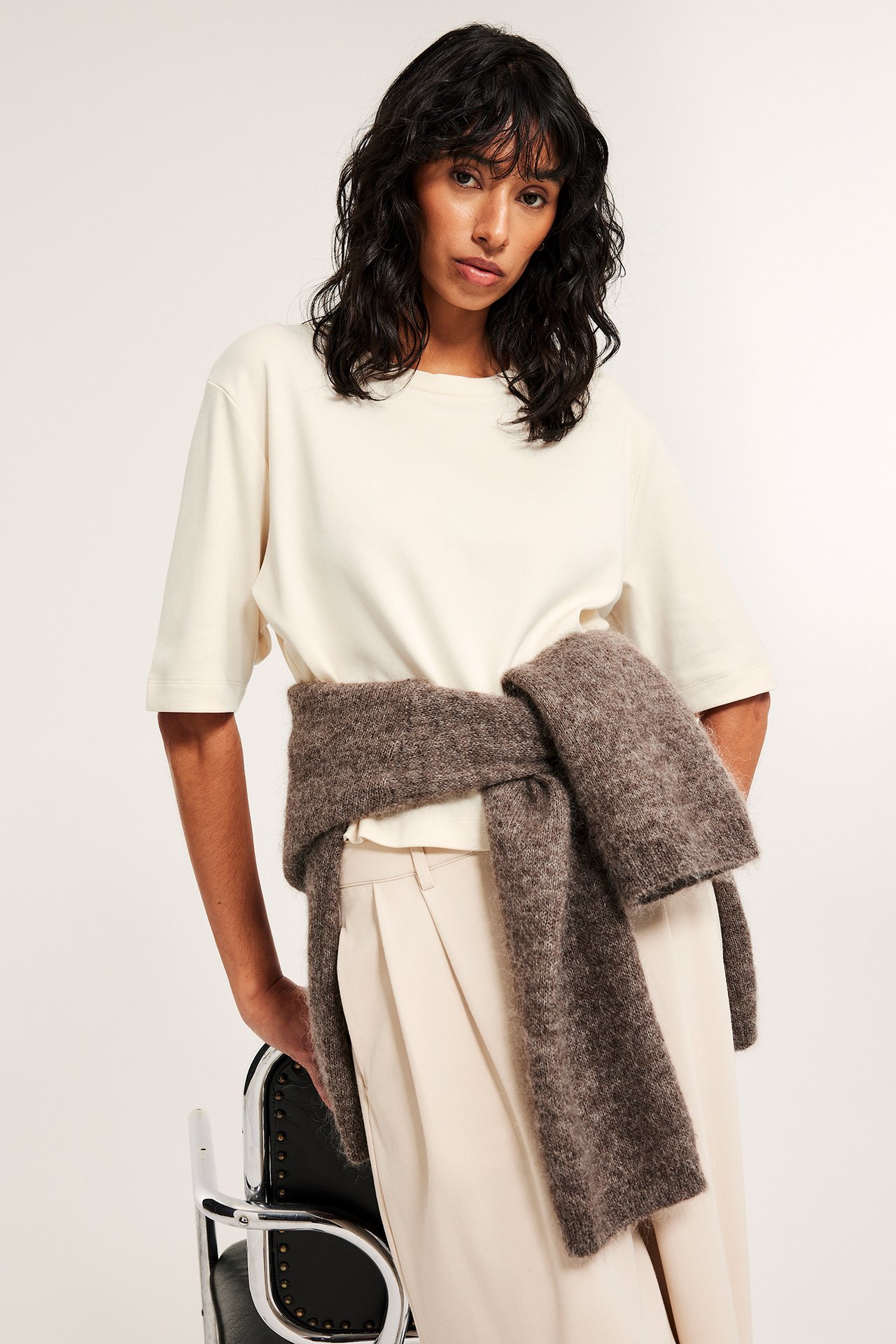 Nainen yllään luonnonvalkoinen lyhyt puuvillainen t-paita, beiget laskostetut housut ja ruskea neule vyötäröllä.