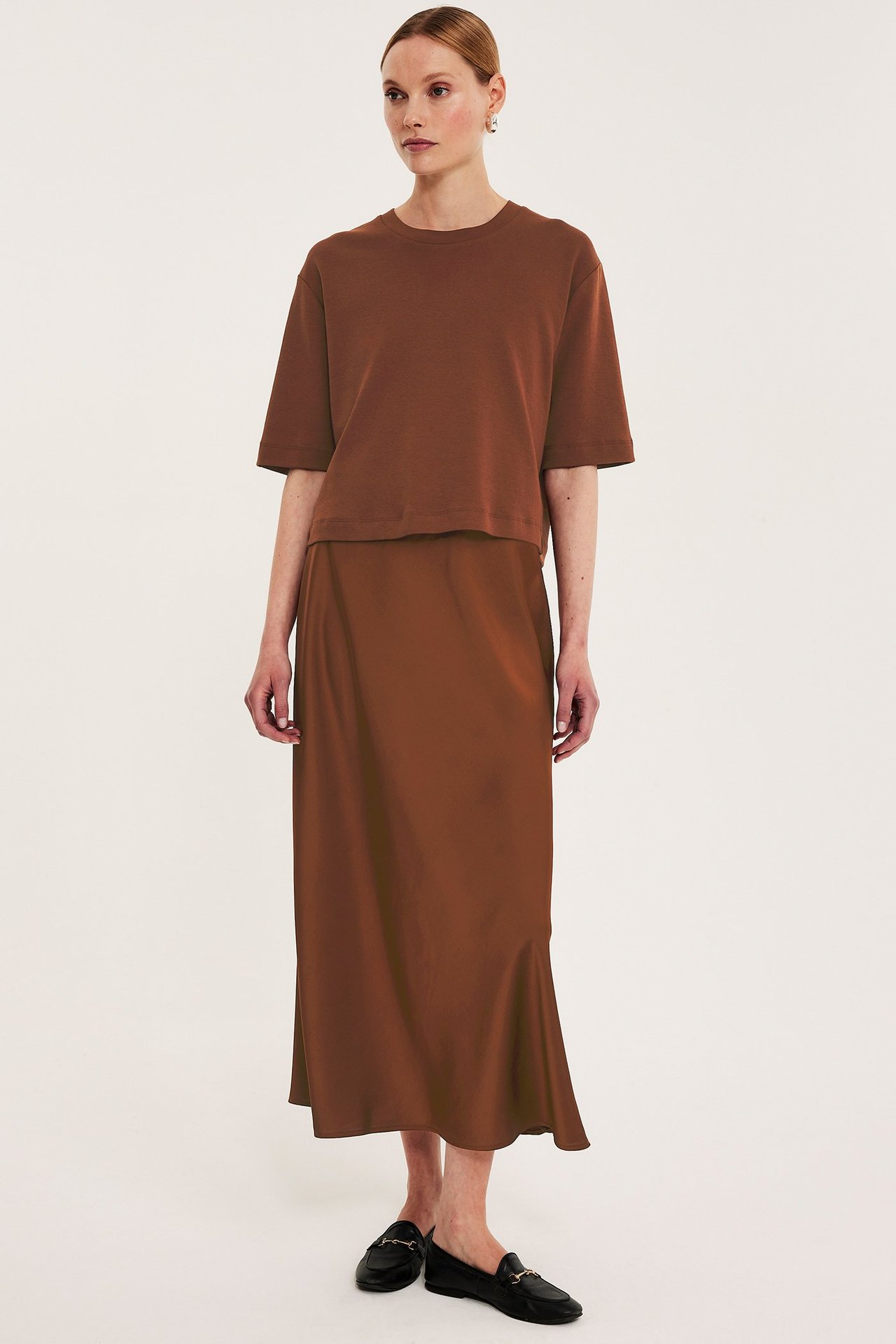 Frontbilde av en kvinne i brun kort bomulls-T-skjorte, brunt midiskjørt og svarte loafers.