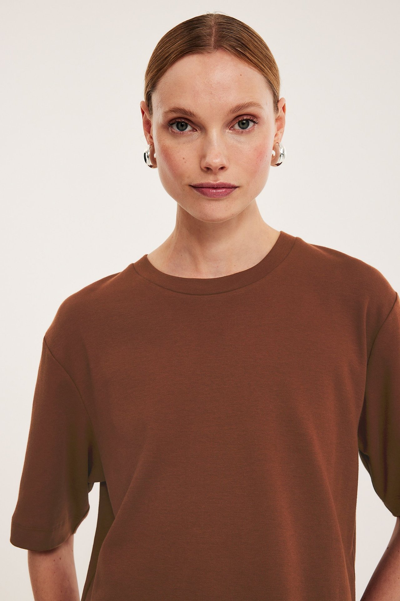 Zbliżenie kobiety w brązowym, krótkim bawełnianym T-shircie i srebrnych kolczykach kołach, widok z przodu.