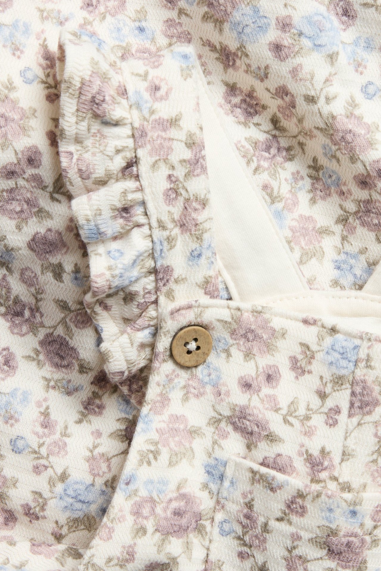 Zbliżenie na ecru kwieciste dżersejowe ogrodniczki niemowlęce z falbankami na szelkach i drewnianym guzikiem.