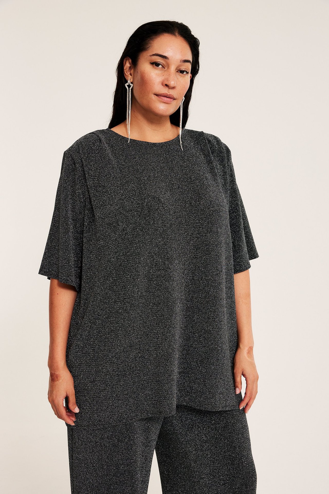 Widok z przodu: kobieta w czarnym, błyszczącym, luźnym topie, dopasowanych spodniach i długich srebrnych kolczykach.