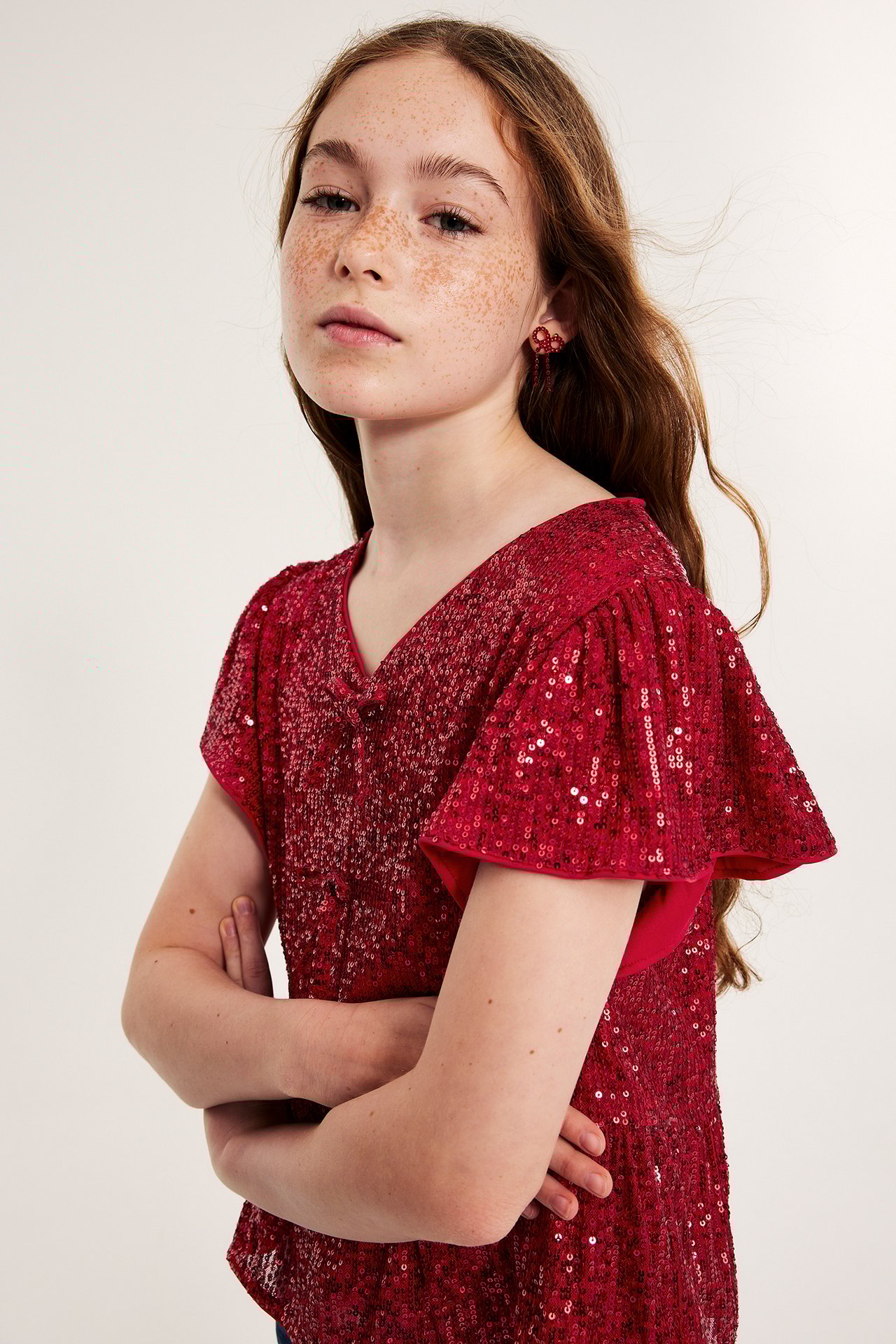 Frontbilde av et barn i rød paljett-V-hals topp med volangermer og røde dinglende øredobber.