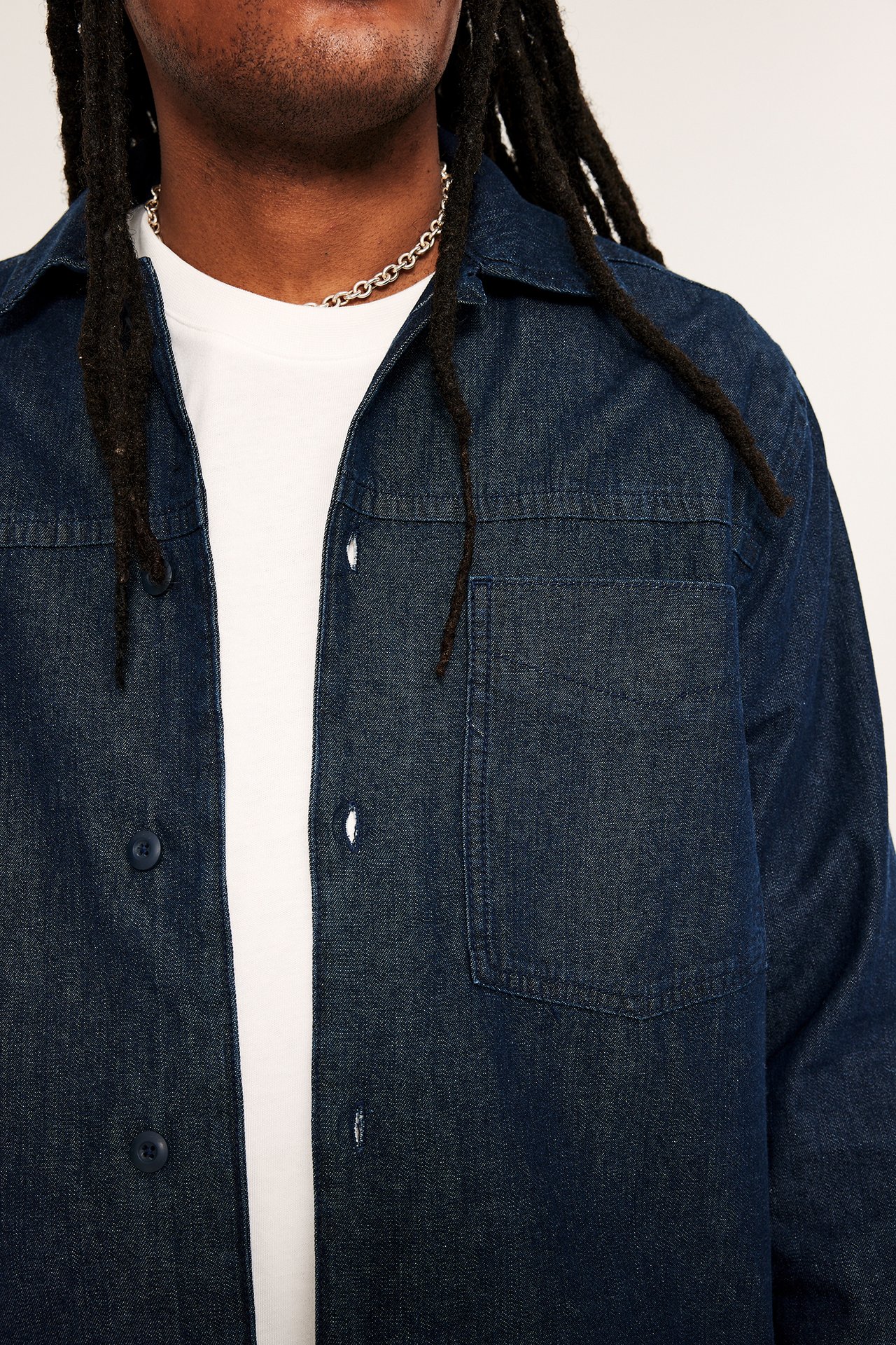 Närbild framifrån: man bär mörk denim overshirt med bröstficka, vit t-shirt, silverhalsband.